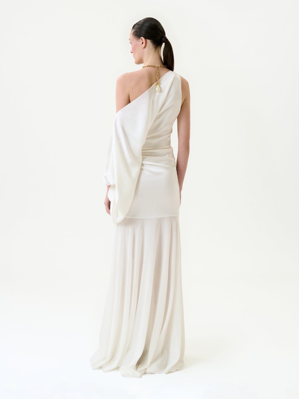 A woman with her dark hair in a ponytail stands against a plain white background, wearing the Emilia Dress—a flowing off-shoulder silk maxi with a single sleeve—and a gold necklace.