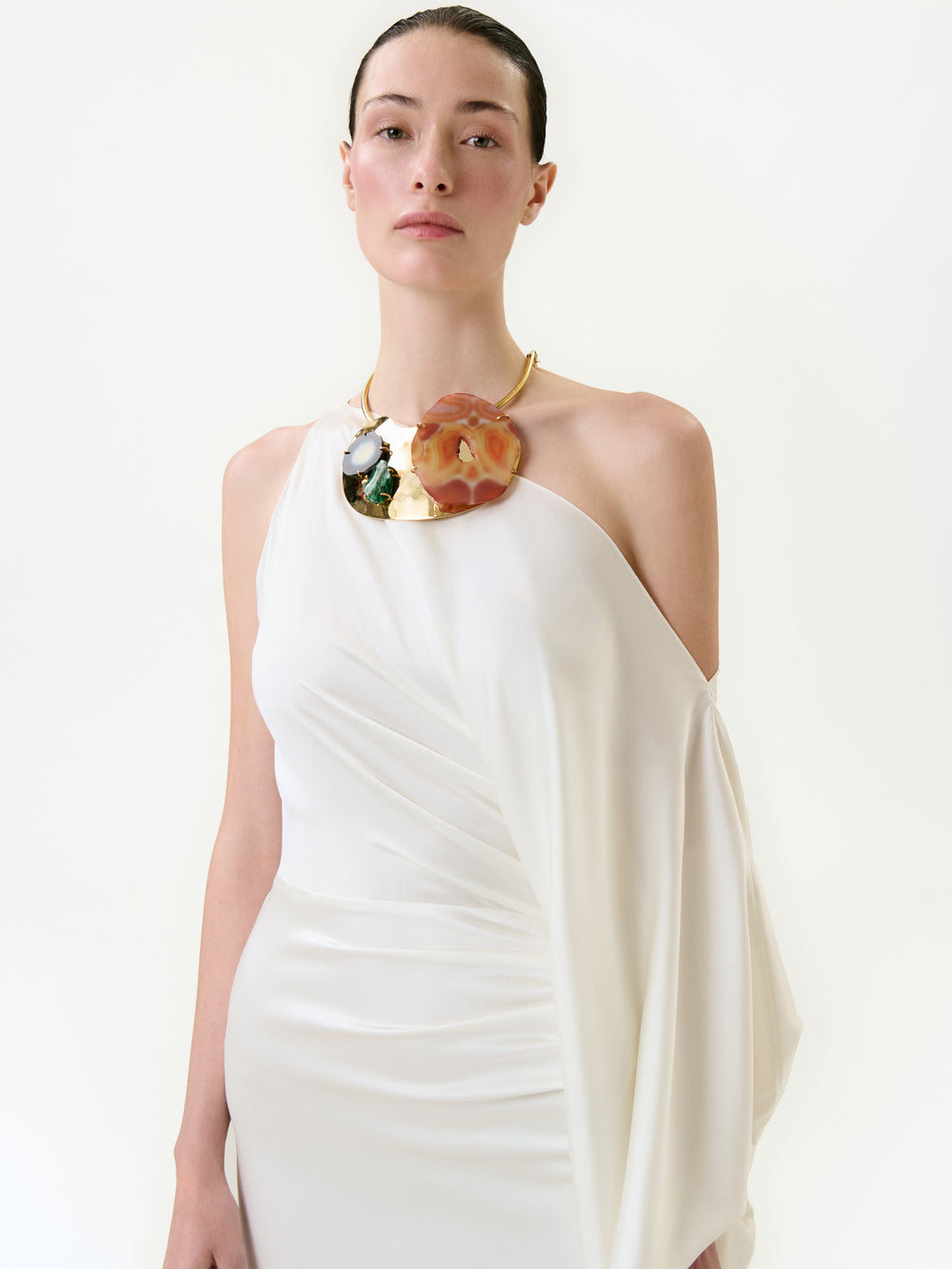 A person models the Emilia Dress, an elegant one-shoulder white maxi crafted from luxurious Italian fabric, paired with a bold statement necklace, against a plain white background.
