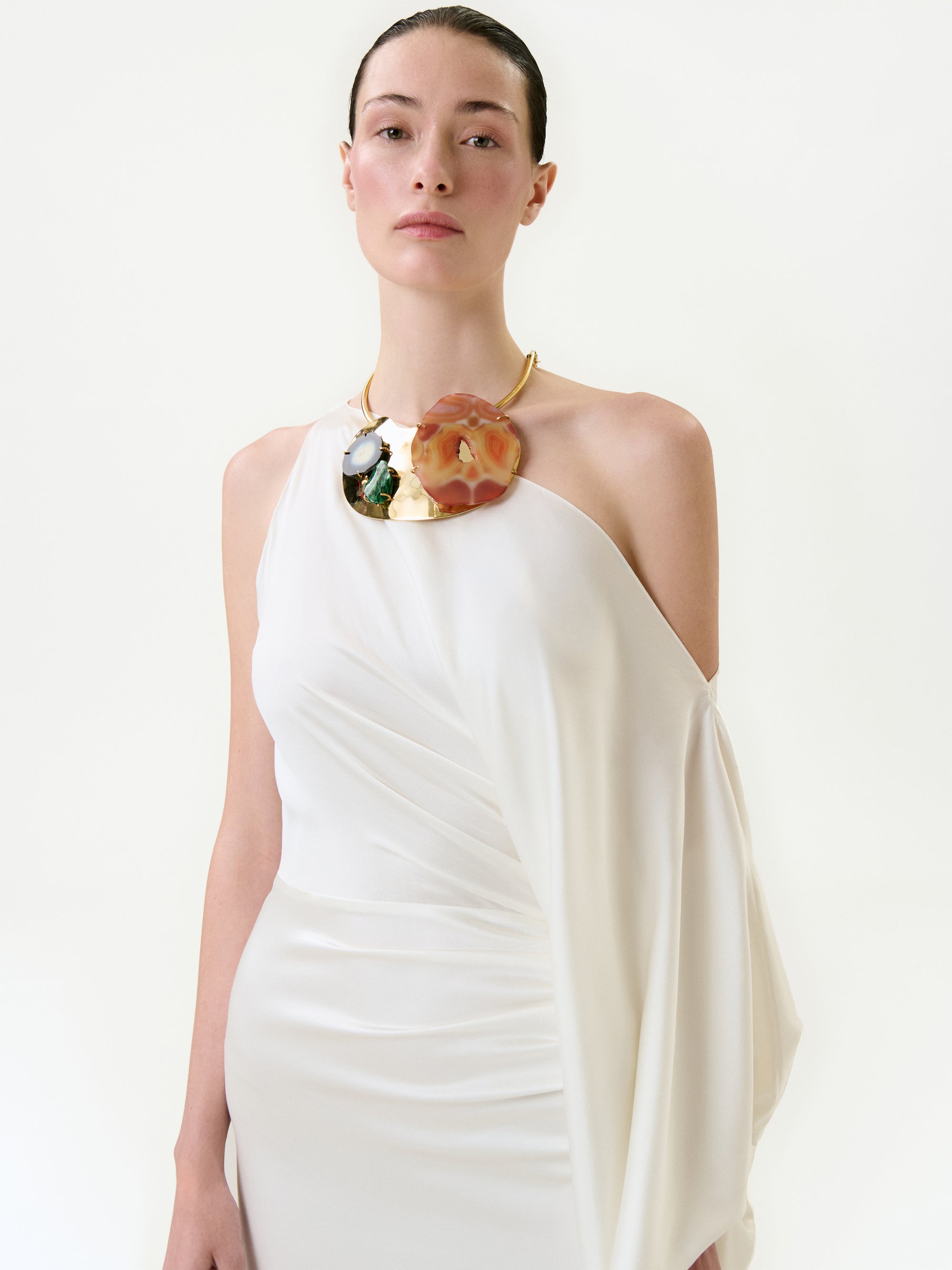 A person models the Emilia Dress, an elegant one-shoulder white maxi crafted from luxurious Italian fabric, paired with a bold statement necklace, against a plain white background.
