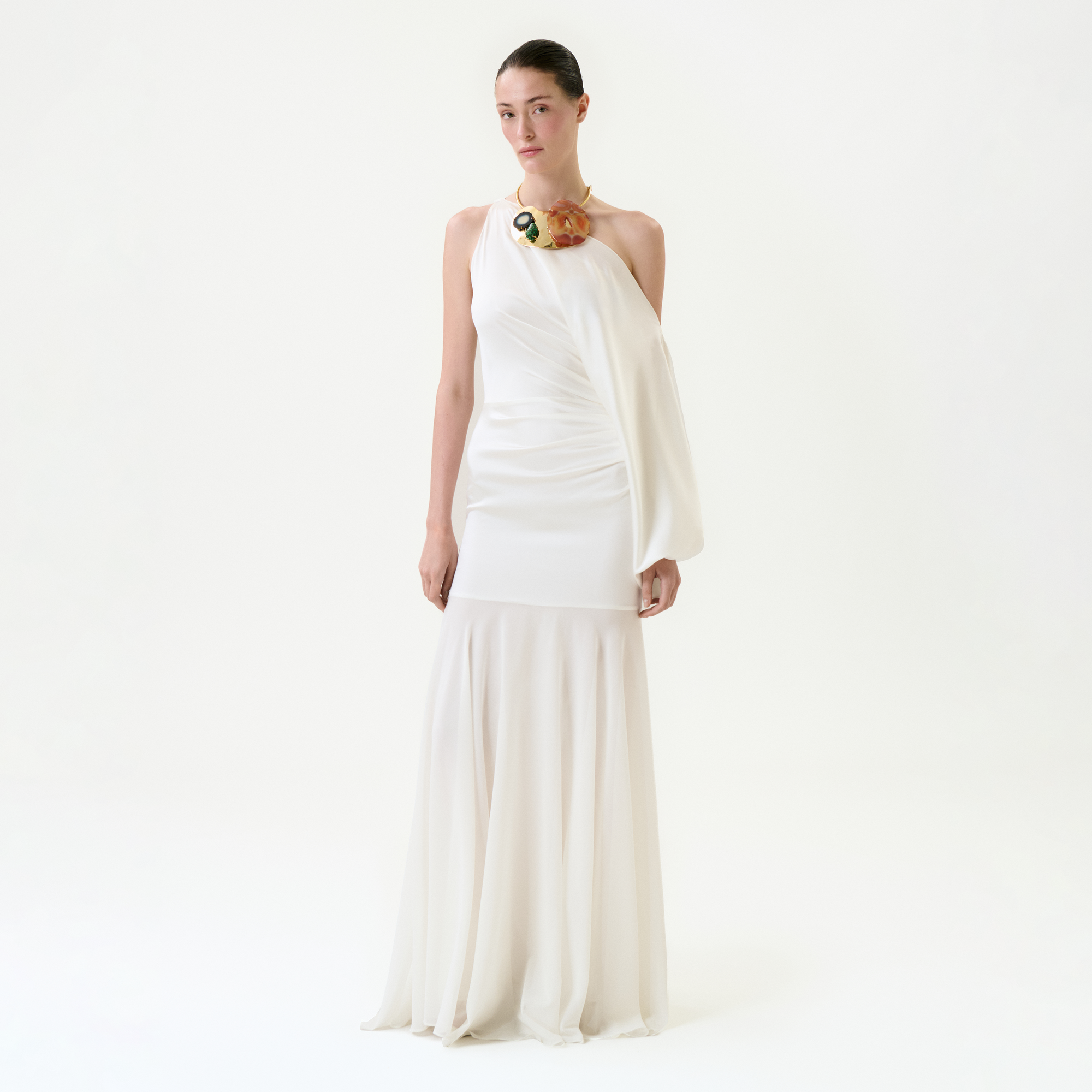 A woman stands against a plain white background wearing the Emilia Dress, a long, elegant one-shoulder maxi crafted from luxurious Italian fabric. Her hair is pulled back and she accessorizes with a bold, colorful floral necklace.