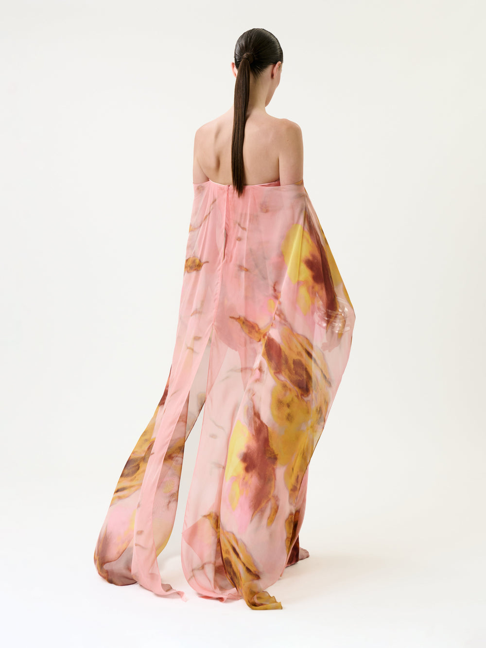 A person with long, dark hair stands barefoot, facing away against a white background, wearing the Lovane Dress—a sheer pink and yellow floral garment.