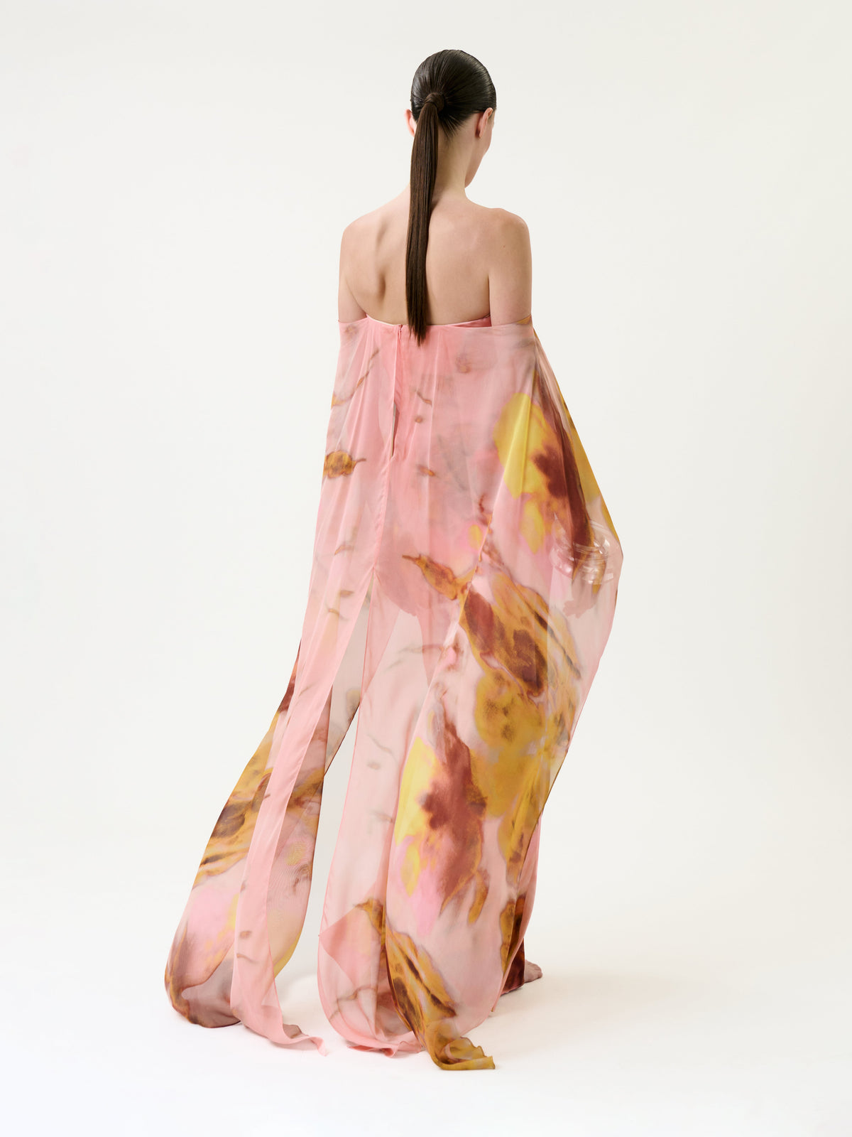 A person with long, dark hair stands barefoot, facing away against a white background, wearing the Lovane Dress—a sheer pink and yellow floral garment.