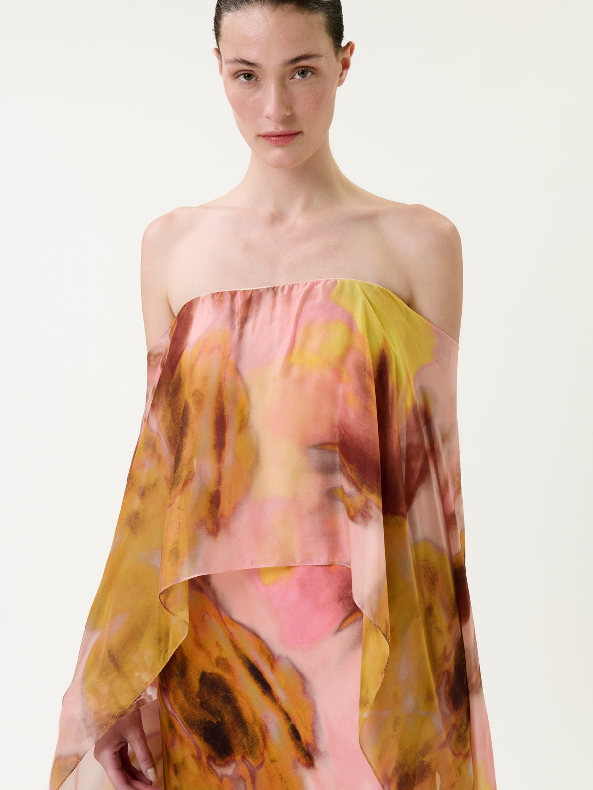 A woman with dark hair pulled back wears the Lovane Dress—a strapless, flowing piece featuring a pink, yellow, and brown abstract pattern—standing against a plain light background.