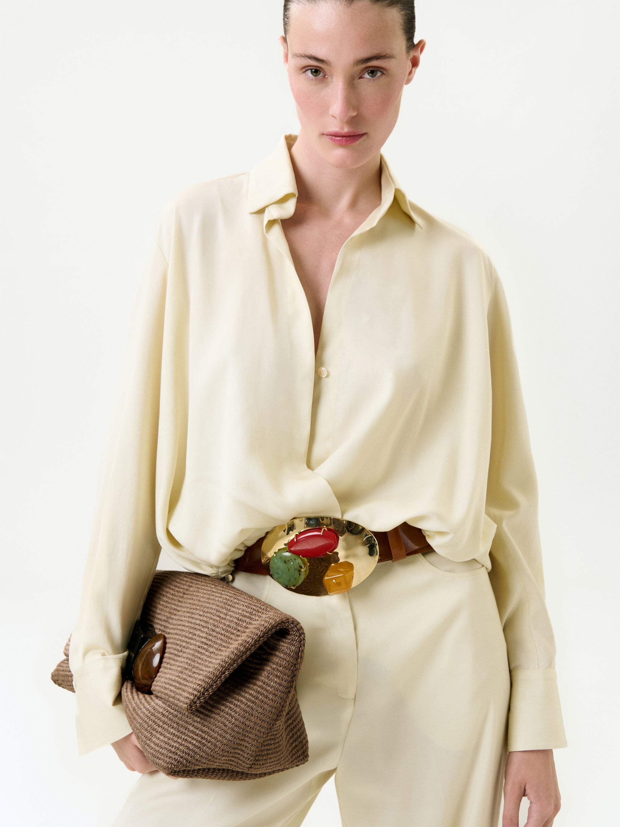 A woman in the loose, cream-colored Saula Blouse and matching Italian-fabric pants holds a textured brown clutch. She wears a bold, colorful statement belt buckle and stands against a plain white background.