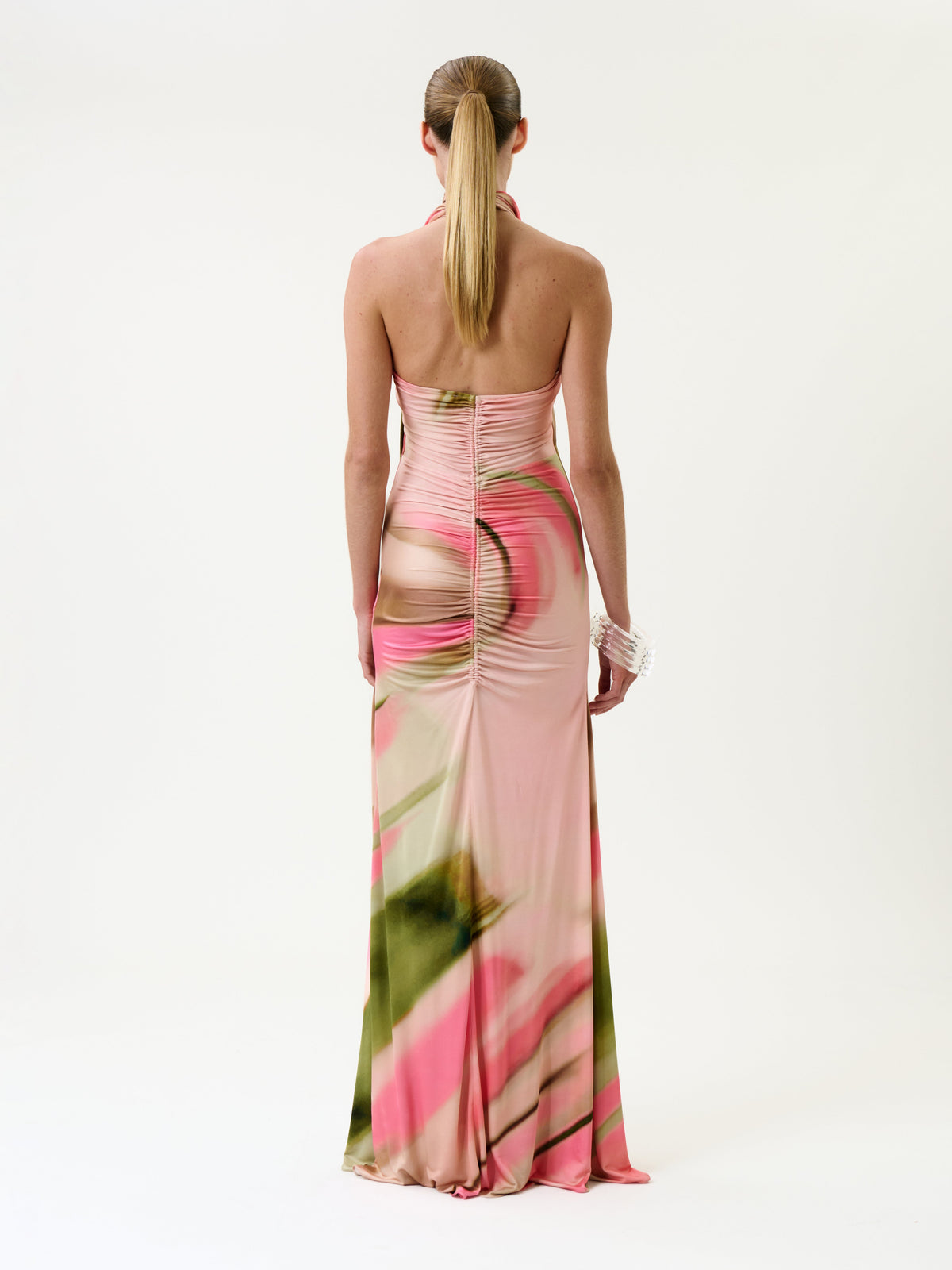 A person with a ponytail stands facing away, wearing the Maika Dress—a pink and green halter-neck gown featuring ruching and cutout details—against a plain white background.