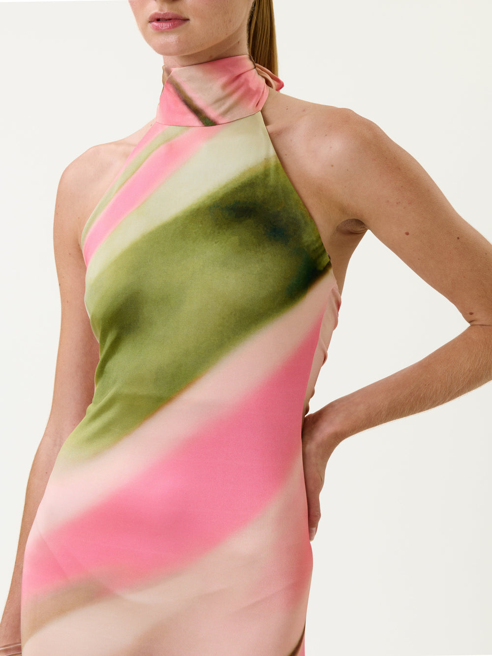 A person wearing the Maika Dress—a sleeveless halter-neck style featuring pink, green, and cream diagonal patterns—stands with one hand on their hip against a plain background.