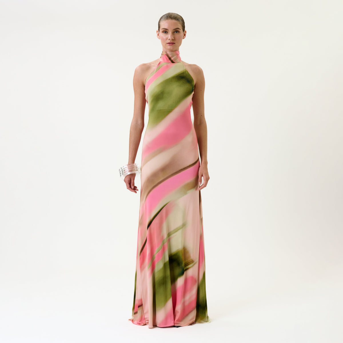 A woman stands against a white background, wearing the Maika Dress—a long, halter-neck dress featuring a pink and green abstract pattern—paired with a chunky white bracelet.