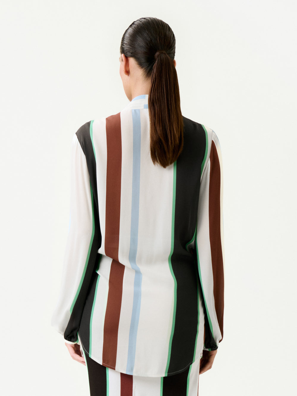 A person with long brown hair in a low ponytail is seen from behind wearing the Honey Blouse—a relaxed fit, long-sleeve top made of 100% viscose with vertical white, brown, green, blue, and black stripes—against a plain white background.