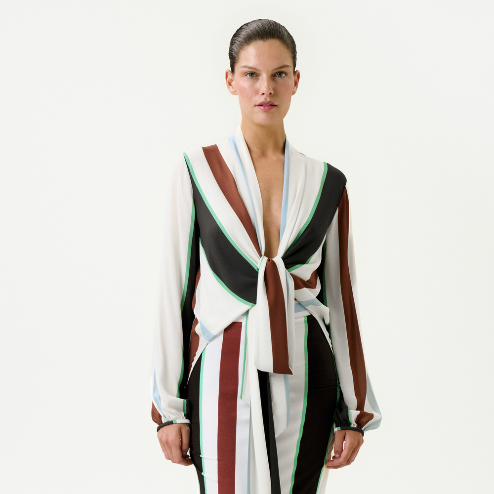 A person stands facing forward, wearing the Honey Blouse—a relaxed fit, long-sleeve top with a deep V-neck and front tie, featuring white, brown, black, and green vertical stripes. Made from 100% viscose. Plain white background.