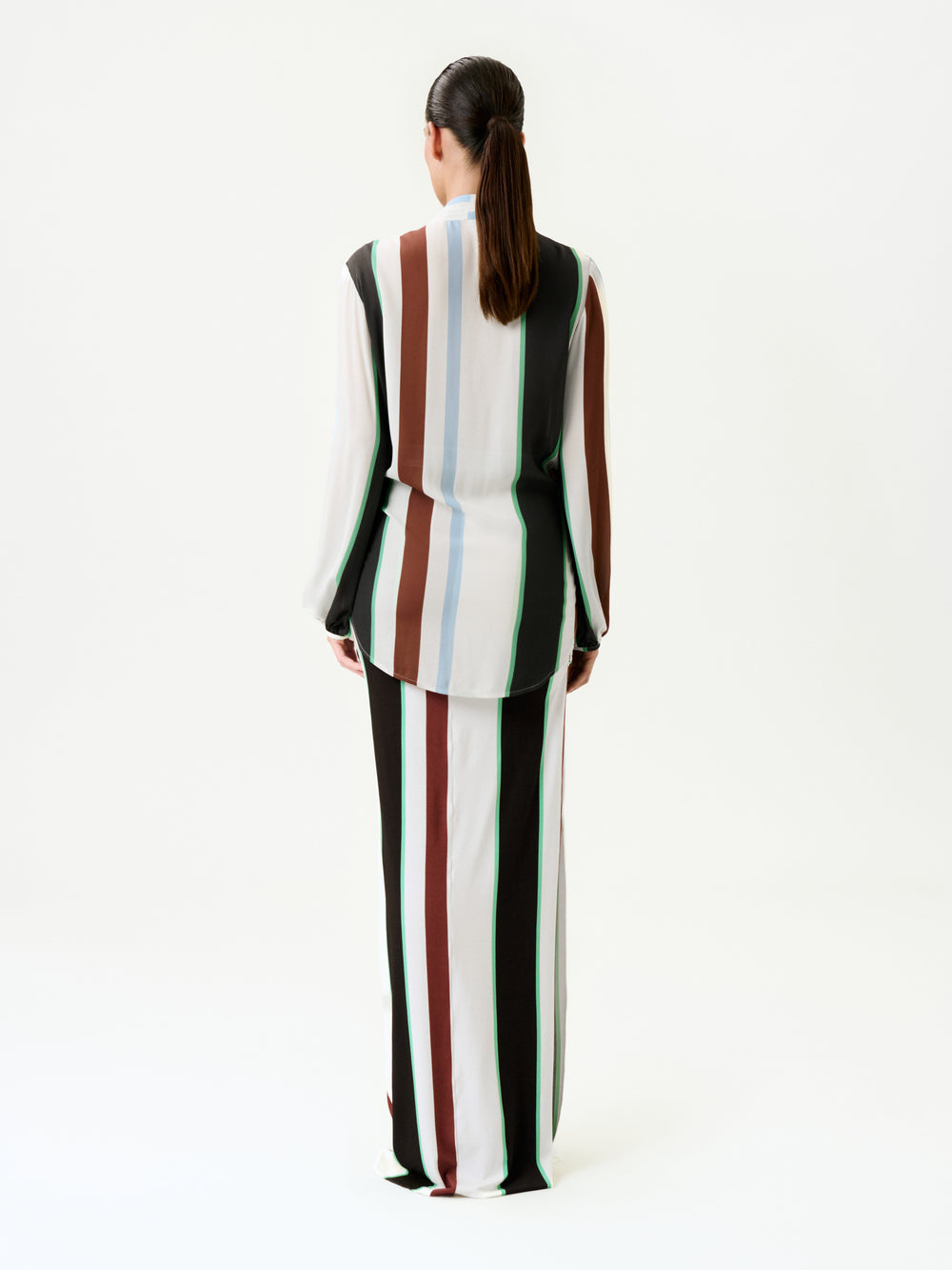 A person with long dark hair in a ponytail stands facing away, wearing a slim fit long-sleeved top and the Toni Skirt with vertical black, white, brown, blue, and green stripes on a plain white background.