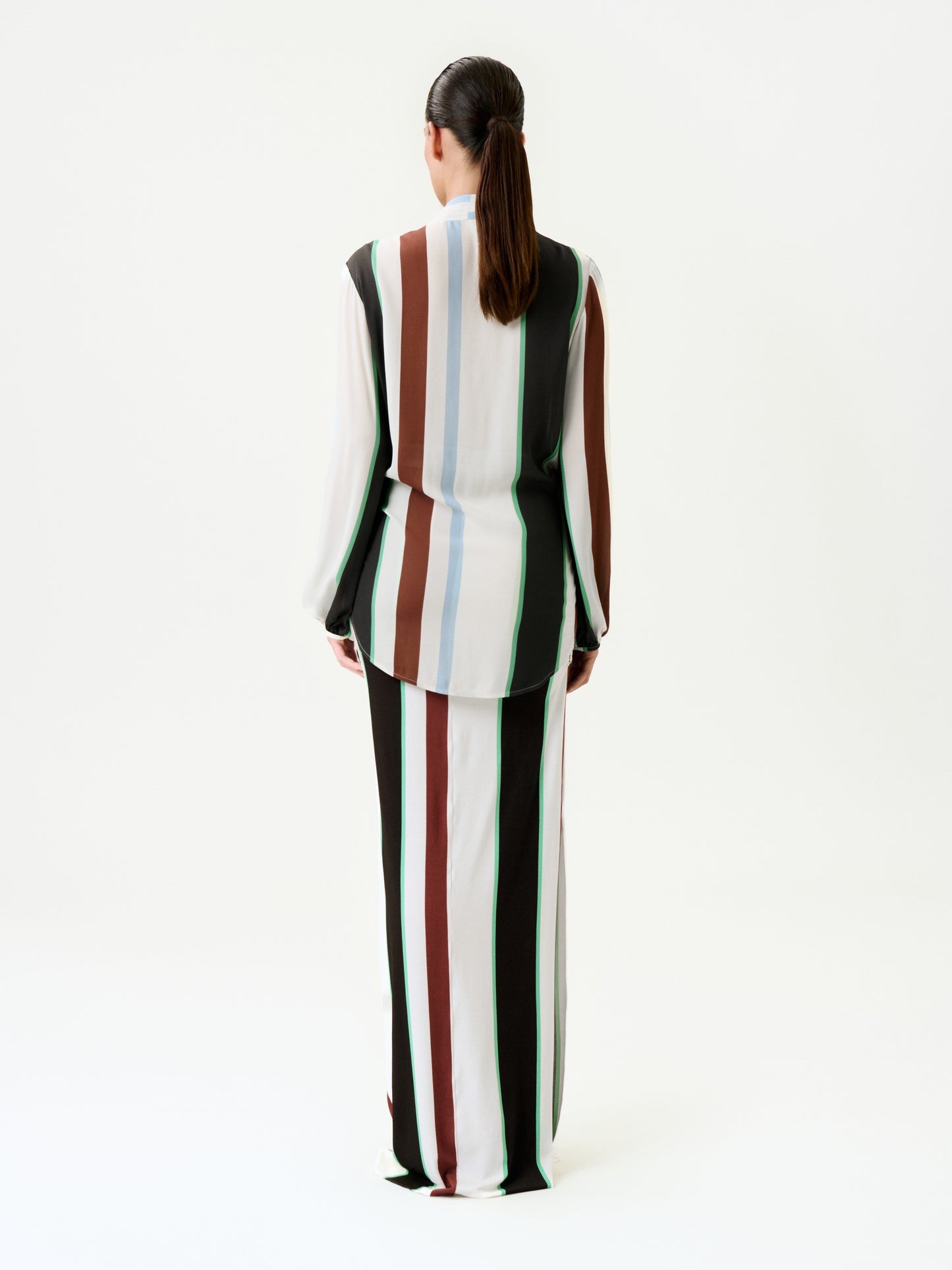 A person with long dark hair in a ponytail stands facing away, wearing a slim fit long-sleeved top and the Toni Skirt with vertical black, white, brown, blue, and green stripes on a plain white background.