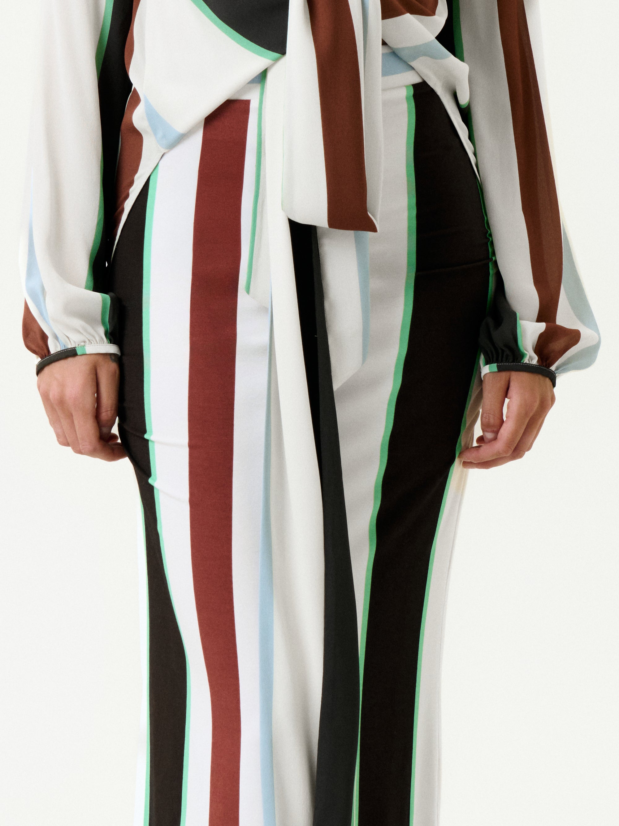 A person wears the Toni Skirt—a flowing, draped skirt with vertical stripes in brown, green, white, light blue, and black. Made in Colombia from Italian fabric, it's styled here with a matching long-sleeve top. Hands are relaxed at the sides.