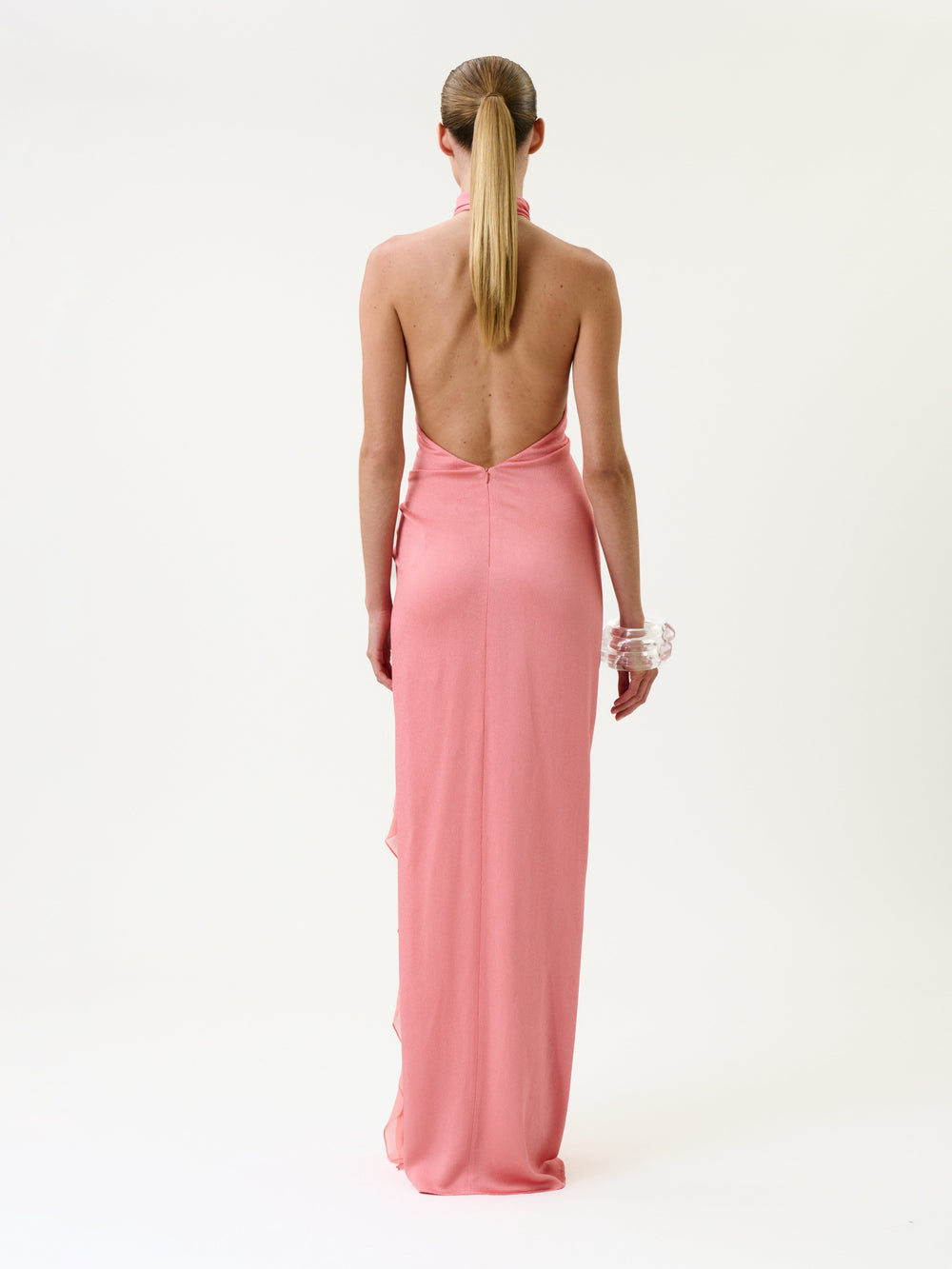 A person with long blond hair in a ponytail wears the Marisela Dress, a backless, floor-length pink gown, along with clear chunky bracelets, standing facing away against a plain white background.