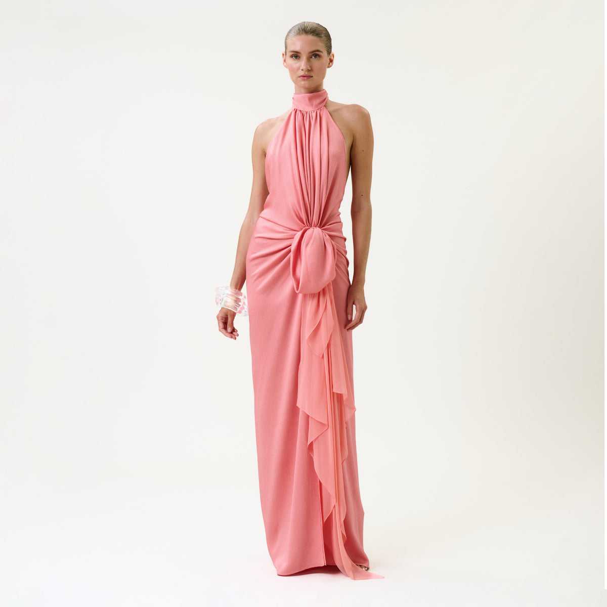 A woman stands wearing the Marisela Dress, a sleeveless, floor-length pink gown featuring a halter neckline, draped front, and gathered knot detail at the waist. She has slicked-back hair, accessorized with a white bracelet, against a plain white background.