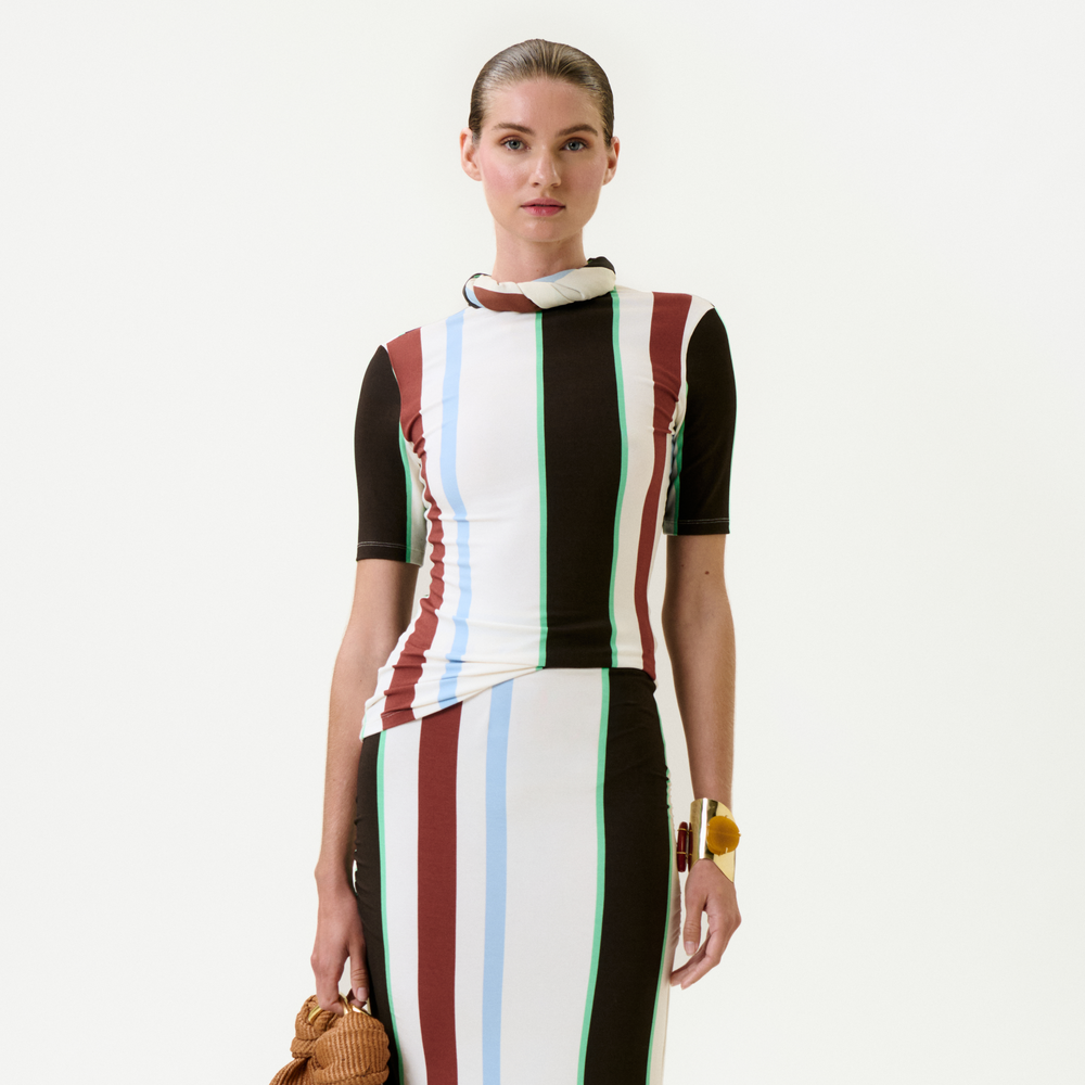 A woman stands against a plain background, wearing the Rowena Top—a slim fit piece with vertical stripes in white, brown, blue, red, and green. She carries a small brown bag, wears a chunky bracelet, and has her hair pulled back.