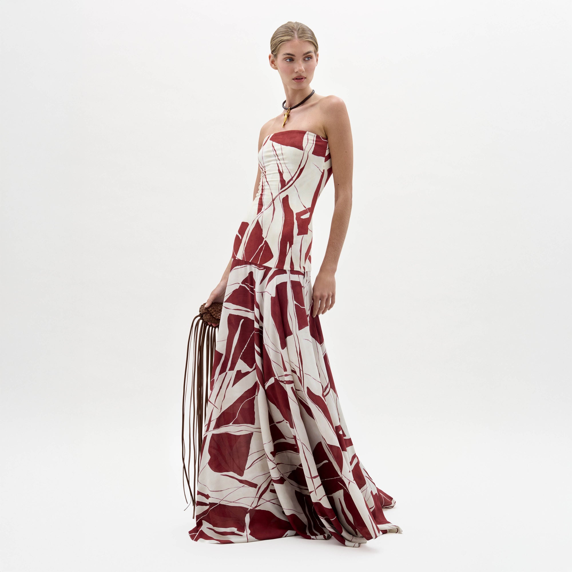 A woman stands wearing the Muriel Dress, a strapless, floor-length gown with a red and white abstract pattern, holding a brown fringe accessory.