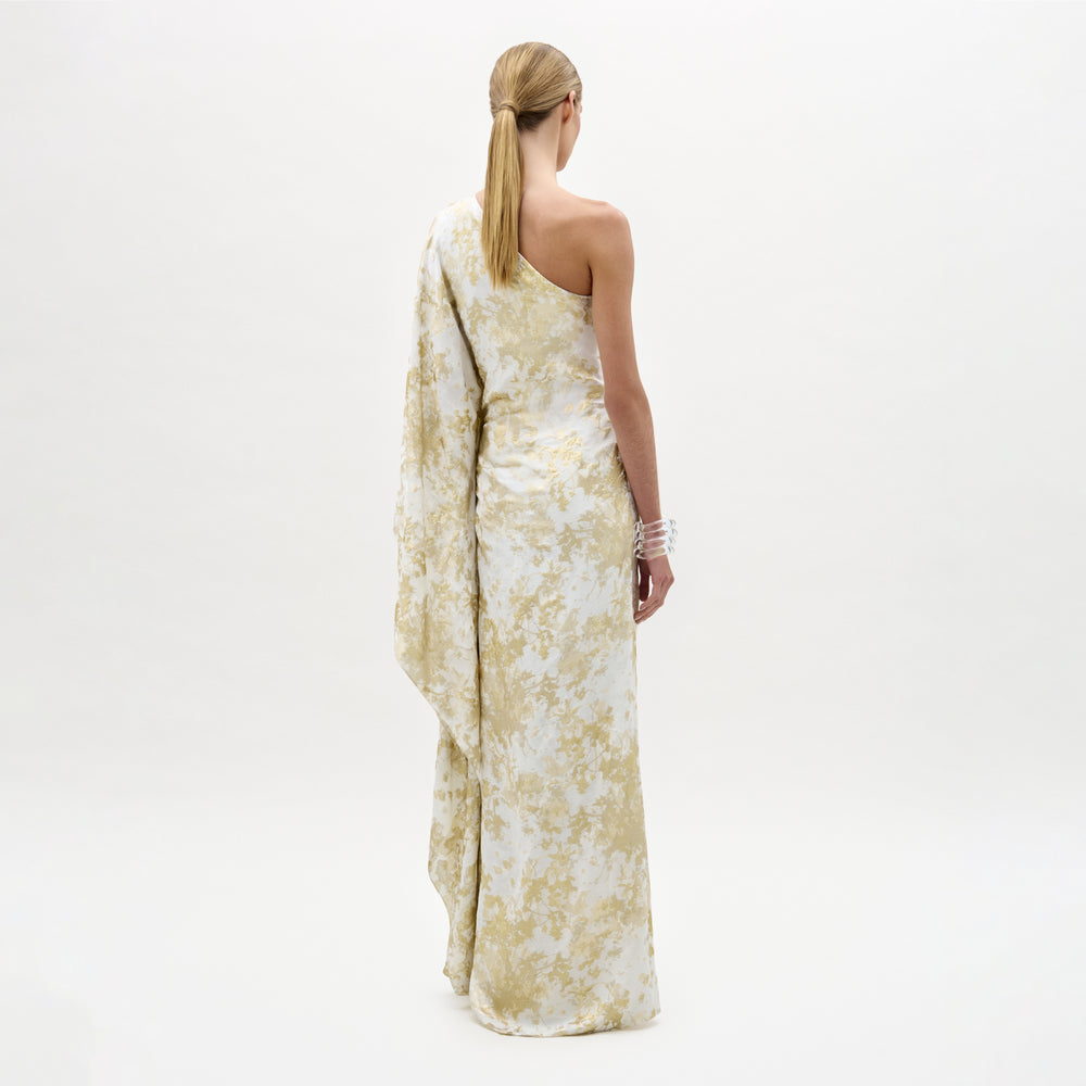 A woman stands facing away, wearing the long, pale yellow and white Afrodita Dress with an asymmetric silhouette and draped sleeve, her blonde hair tied back against a plain white background.