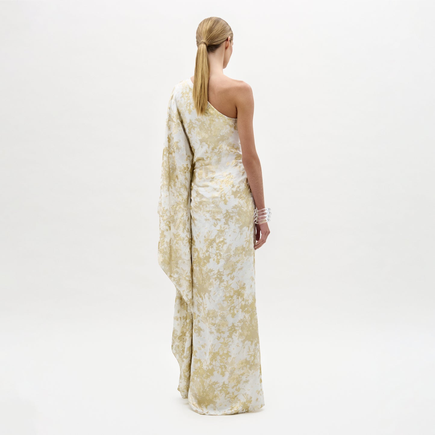 A woman stands facing away, wearing the long, pale yellow and white Afrodita Dress with an asymmetric silhouette and draped sleeve, her blonde hair tied back against a plain white background.
