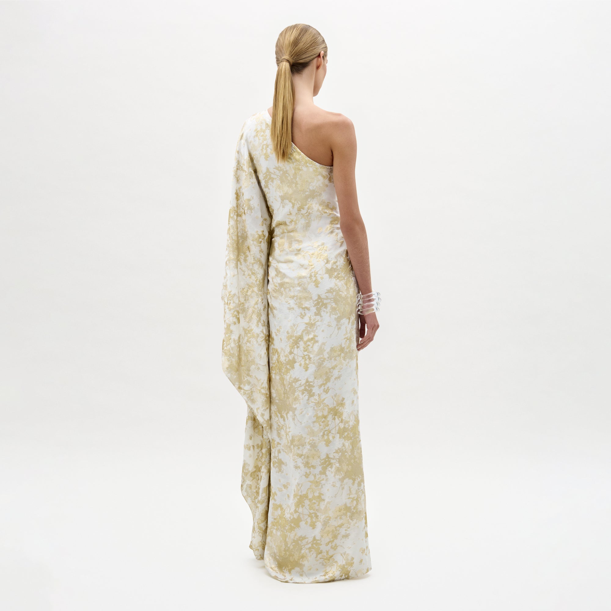 A woman stands facing away, wearing the long, pale yellow and white Afrodita Dress with an asymmetric silhouette and draped sleeve, her blonde hair tied back against a plain white background.