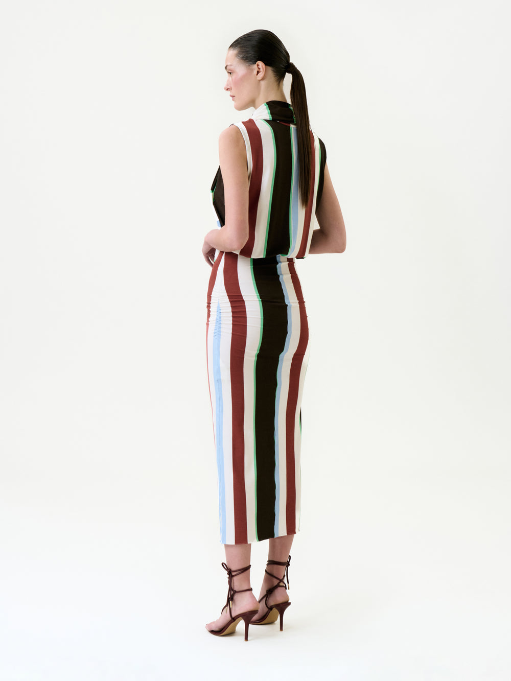 A woman with dark hair in a sleek ponytail stands facing away, wearing the Zaz Dress—a sleeveless, collared midi dress with vertical stripes in brown, white, blue, and maroon—crafted from Italian fabric in Colombia. She pairs it with brown strappy heels.