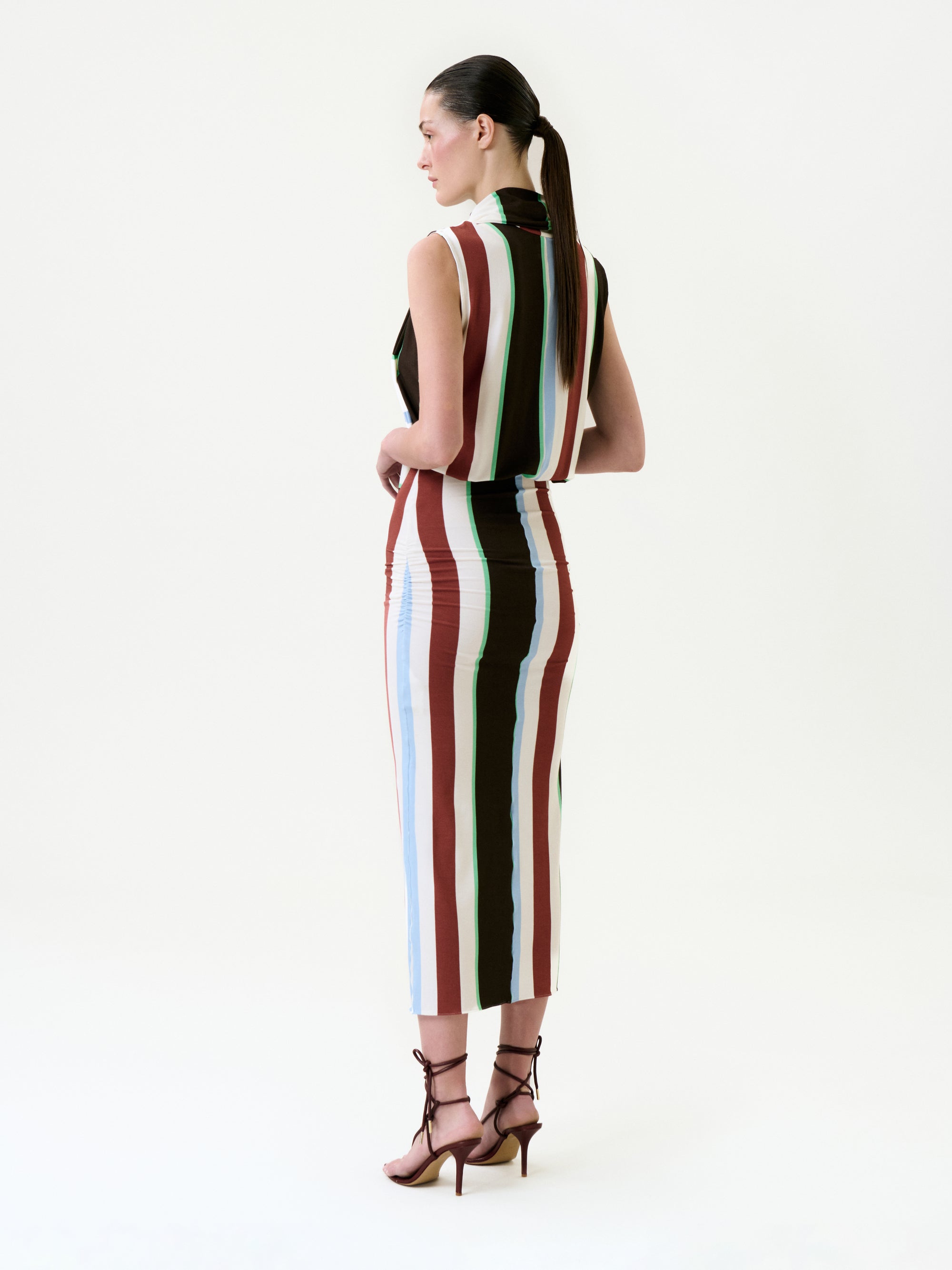 A woman with dark hair in a sleek ponytail stands facing away, wearing the Zaz Dress—a sleeveless, collared midi dress with vertical stripes in brown, white, blue, and maroon—crafted from Italian fabric in Colombia. She pairs it with brown strappy heels.