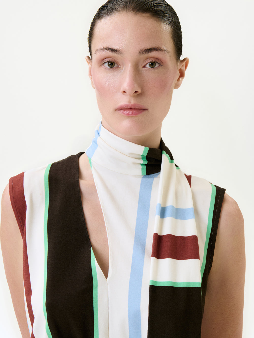 A woman with slicked-back dark hair wears the Zaz Dress, a sleeveless style crafted from Italian fabric, featuring vertical stripes in brown, white, green, blue, and red. She faces the camera with a neutral expression against a plain background.
