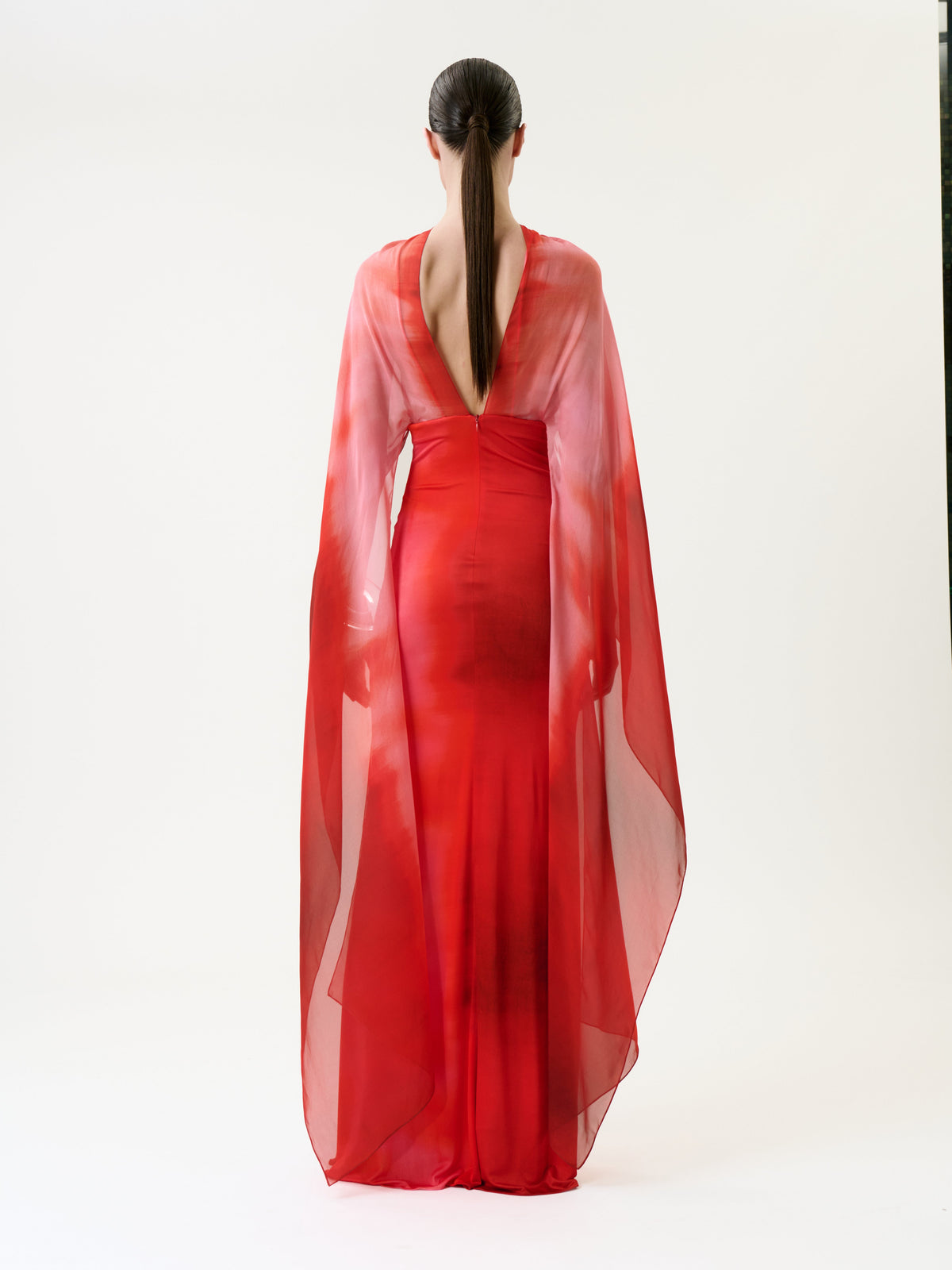 A person with long, dark hair in a low ponytail models the Kendra Dress—a sheer, flowing red gown featuring a deep V-back—standing against a plain white background.