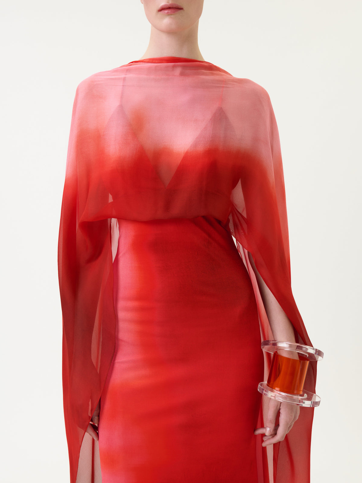 A person wearing the Kendra Dress, a sheer red ombré design featuring cape-like details, is holding a chunky transparent bracelet with an amber accent. The image is cropped at the shoulders and mid-thigh, set against a plain background.