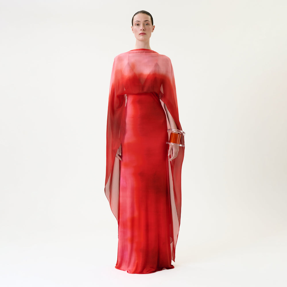 A person wearing the Kendra Dress—a long, sheer red dress with flowing sleeves—stands against a plain white background, holding a round, transparent handbag.
