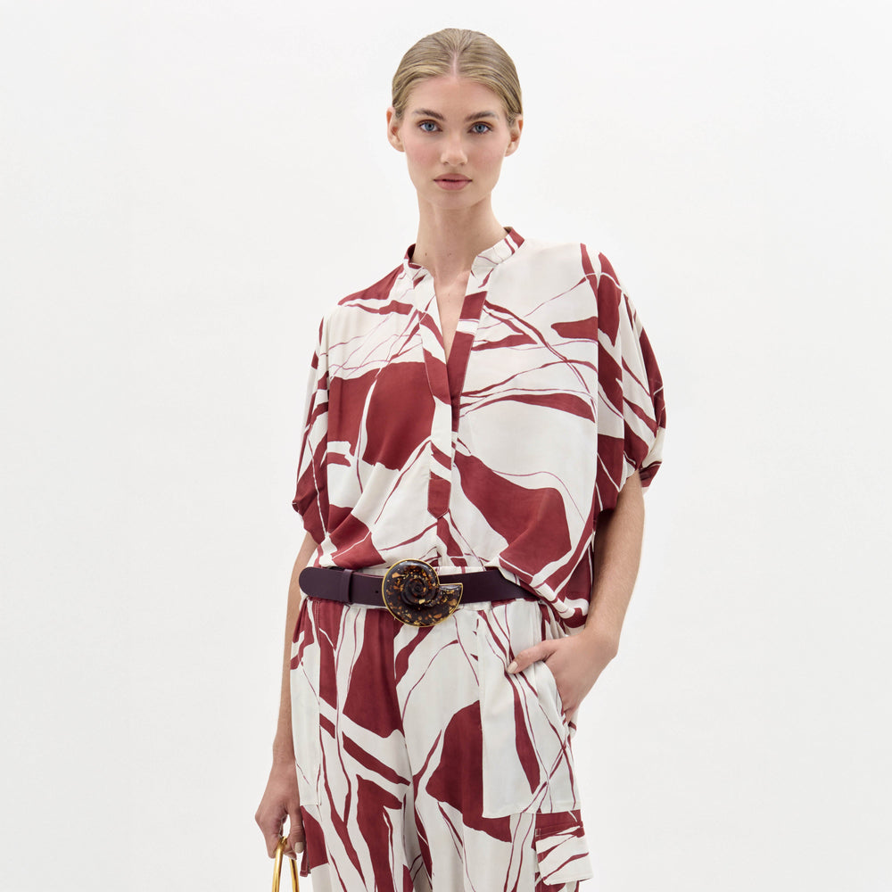A woman poses against a plain white background wearing the Susanne Blouse, featuring a red and white abstract pattern, matched with coordinating pants and accessorized with a black belt with a gold buckle.