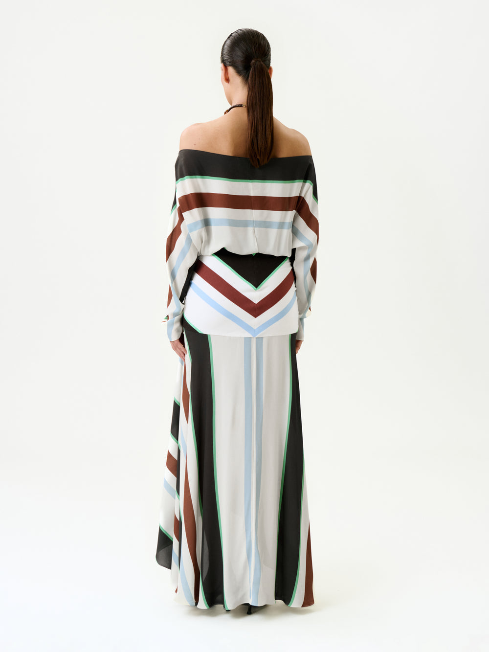 A woman with long dark hair in a low ponytail stands facing away, wearing the Sara Dress—a long, off-the-shoulder maxi dress with wide white, brown, black, blue, and green stripes—made in Colombia.
