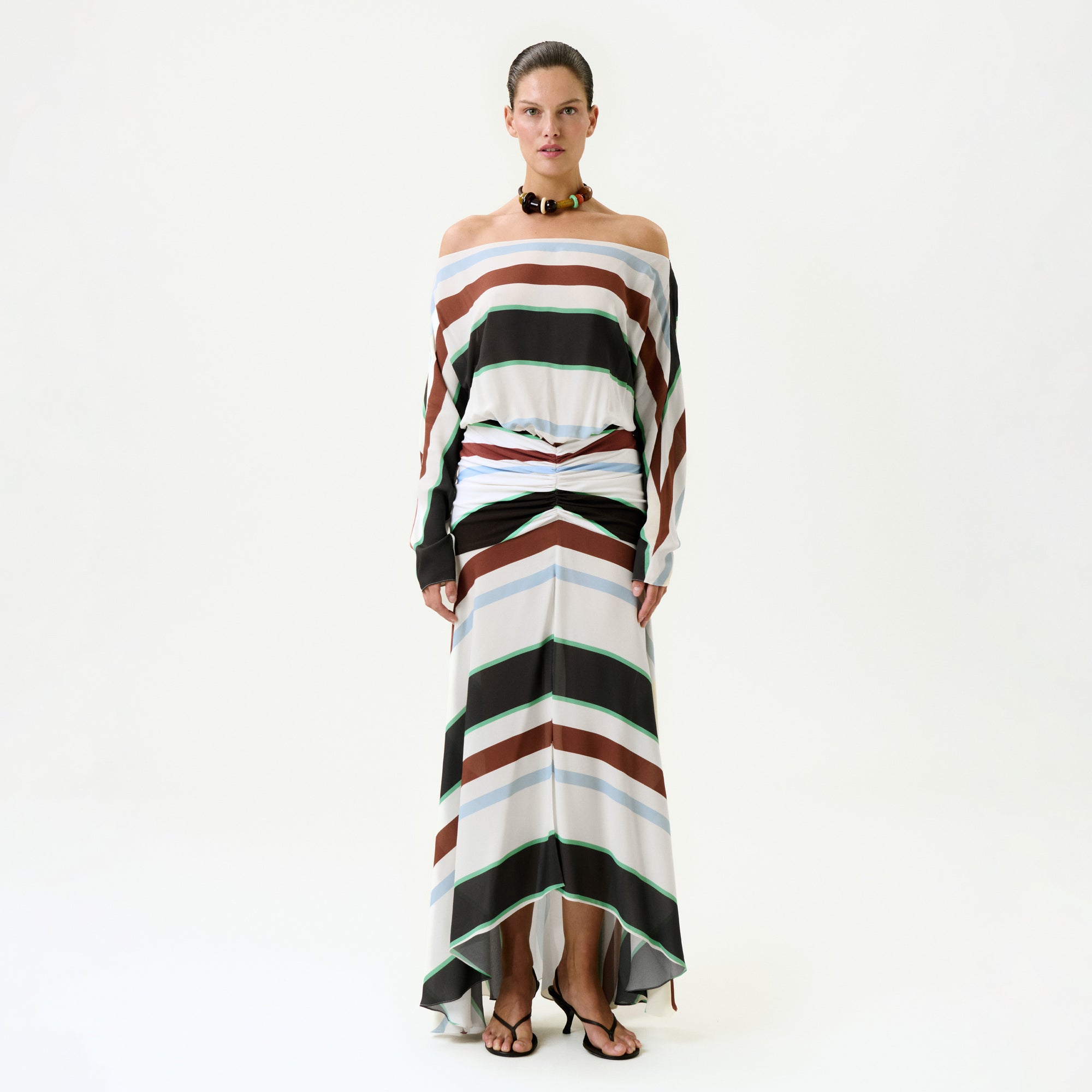 A woman wears the Sara Dress—an off-the-shoulder, long-sleeved maxi with bold brown, black, white, and green horizontal stripes—made in Colombia. She pairs it with black sandals and a chunky necklace against a white background.