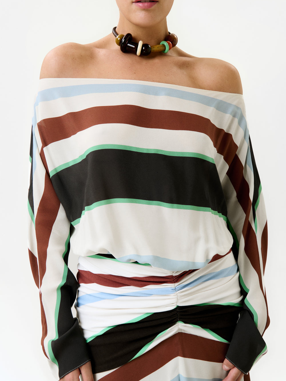 A person wears the Sara Dress—an off-shoulder maxi crafted in Colombia from premium Italian fabric, featuring diagonal stripes in brown, black, green, and blue—paired with a chunky beaded necklace. The background is plain white.