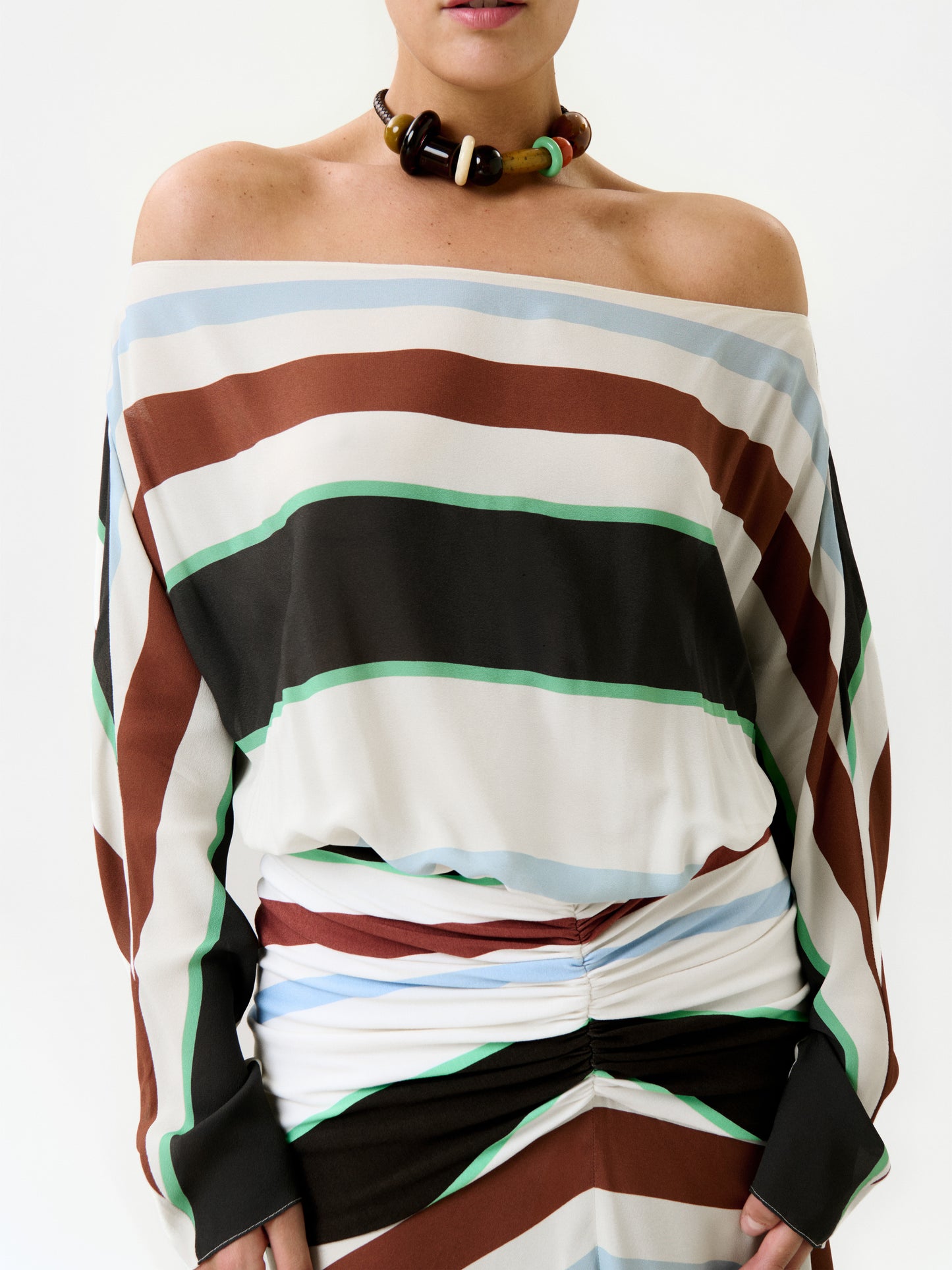 A person wears the Sara Dress—an off-shoulder maxi crafted in Colombia from premium Italian fabric, featuring diagonal stripes in brown, black, green, and blue—paired with a chunky beaded necklace. The background is plain white.
