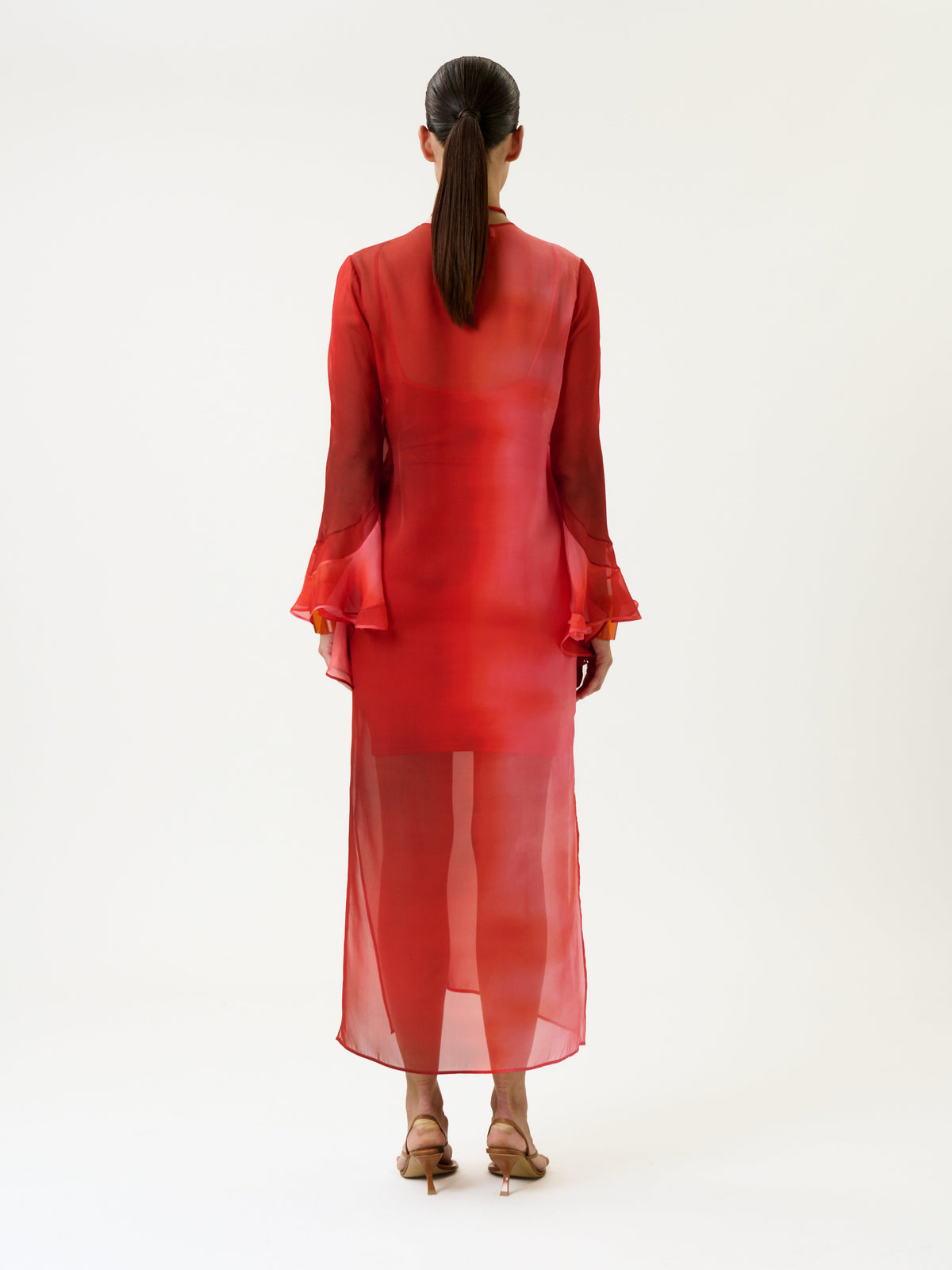A woman with straight brown hair in a ponytail wears the Aries Dress—a sheer, long red dress with flared sleeves—paired with heeled sandals, standing against a plain white background and facing away from the camera.