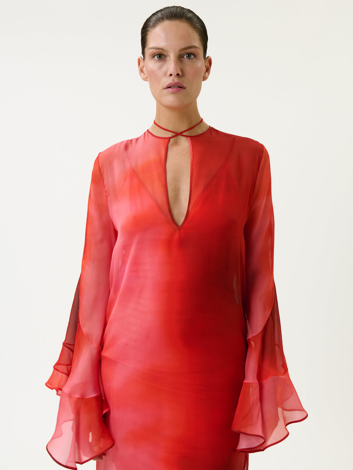 A person wearing the Aries Dress—a sheer, long-sleeve red dress featuring ruffled cuffs and a keyhole neckline—stands against a plain white background, looking directly at the camera.