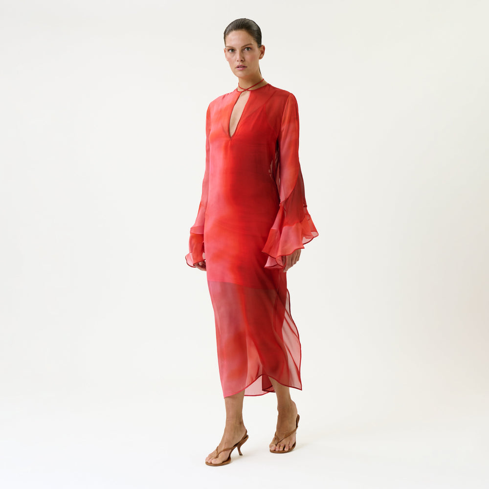 A person wearing the Aries Dress—a sheer, long-sleeved red dress with flared cuffs and a V-neckline—stands barefoot against a plain white background.