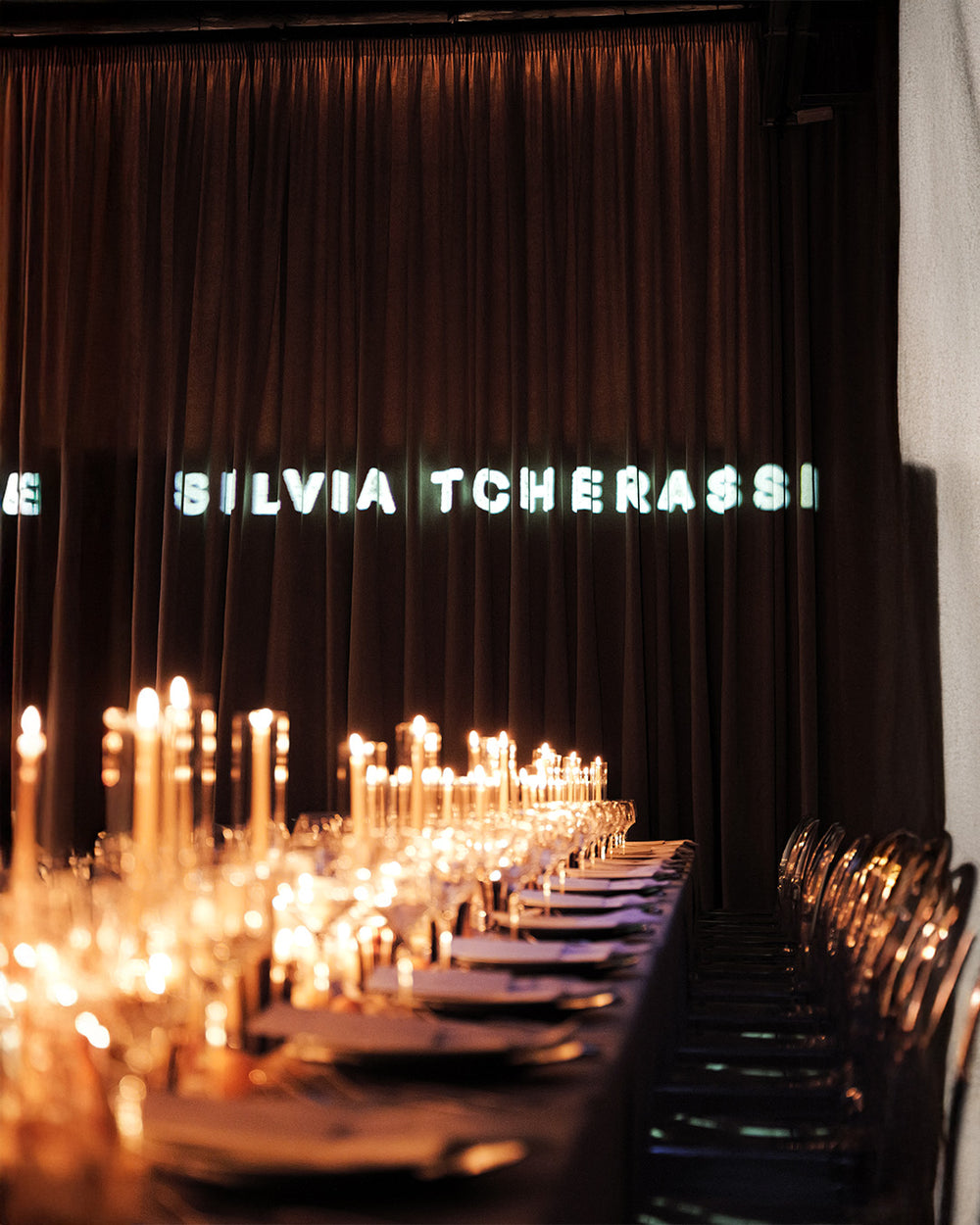 A long banquet table set with lit taper candles, plates, and glassware in front of a dark curtain backdrop with the name 'SILVIA TCHERASSI' illuminated in the background.