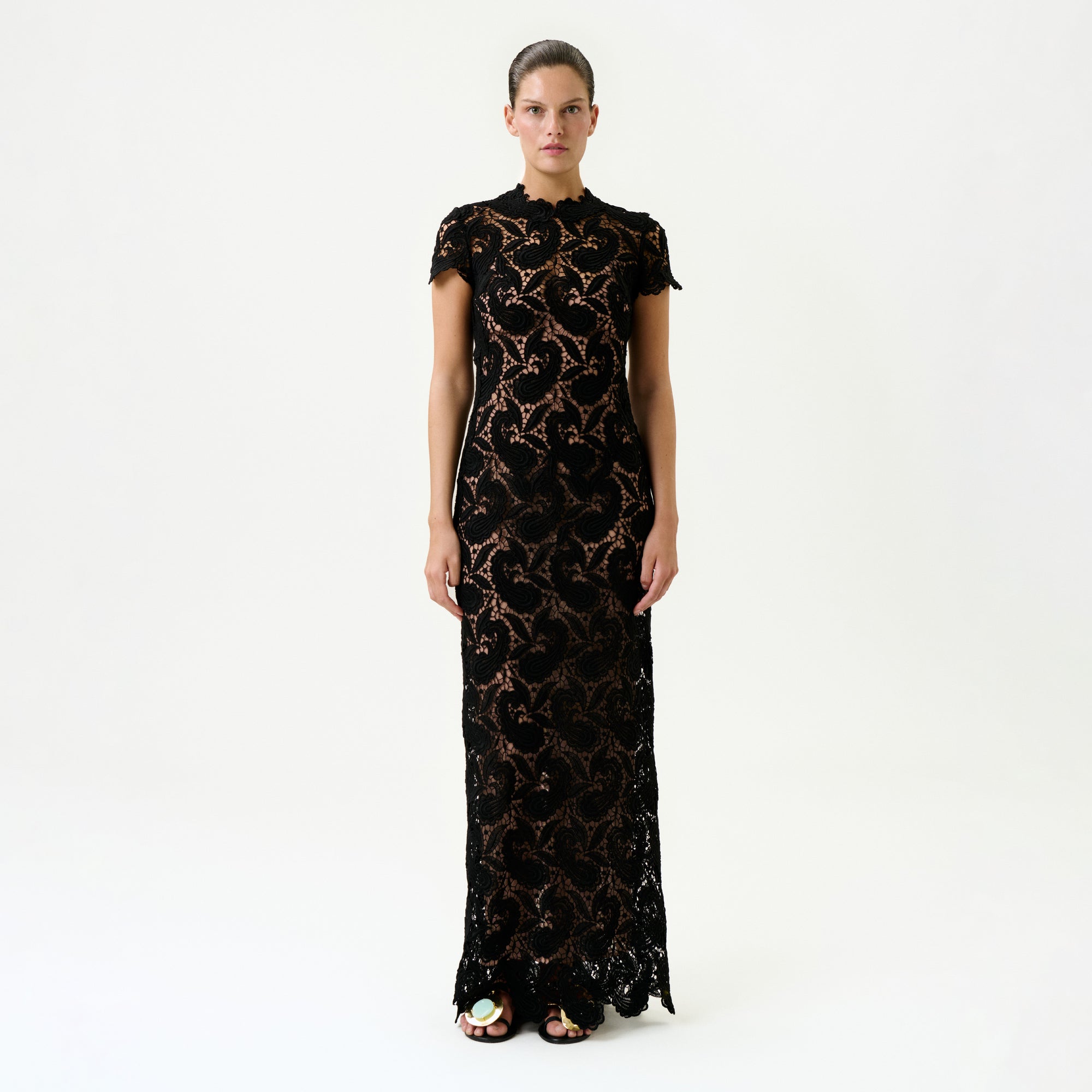 A person stands against a plain white background, wearing the Roxana Dress—a floor-length black lace maxi with a floral pattern, nude underlay, and luxurious Italian fabric—paired with dark sandals.