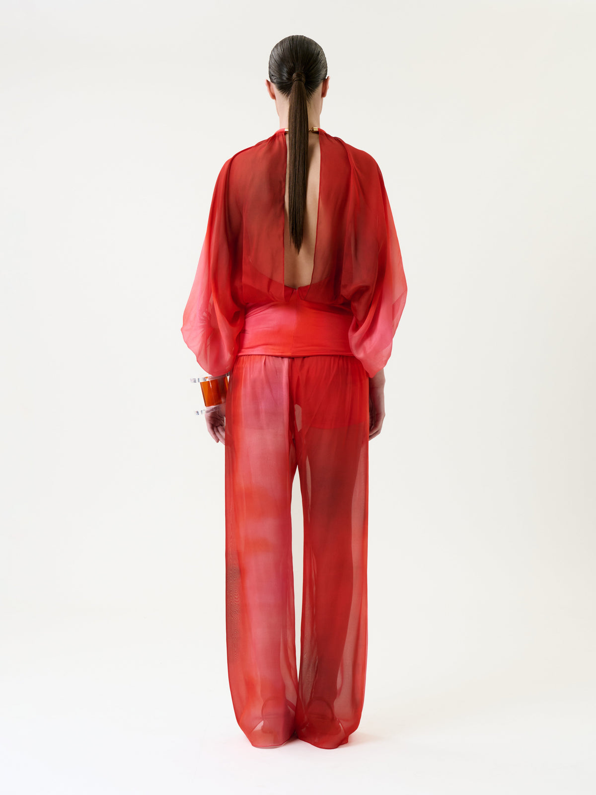 A person with long, straight hair models the Lene Pant in a sheer red ensemble with wide sleeves and pants, standing against a plain white background and facing away from the camera.