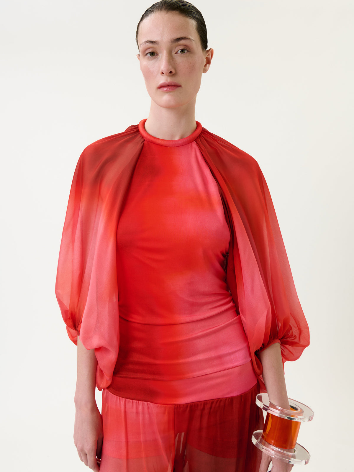 A woman wearing the Lene Pant in a sheer red outfit with loose sleeves stands against a plain white background, holding a clear cylindrical object in her right hand. Her hair is pulled back and her expression is neutral.