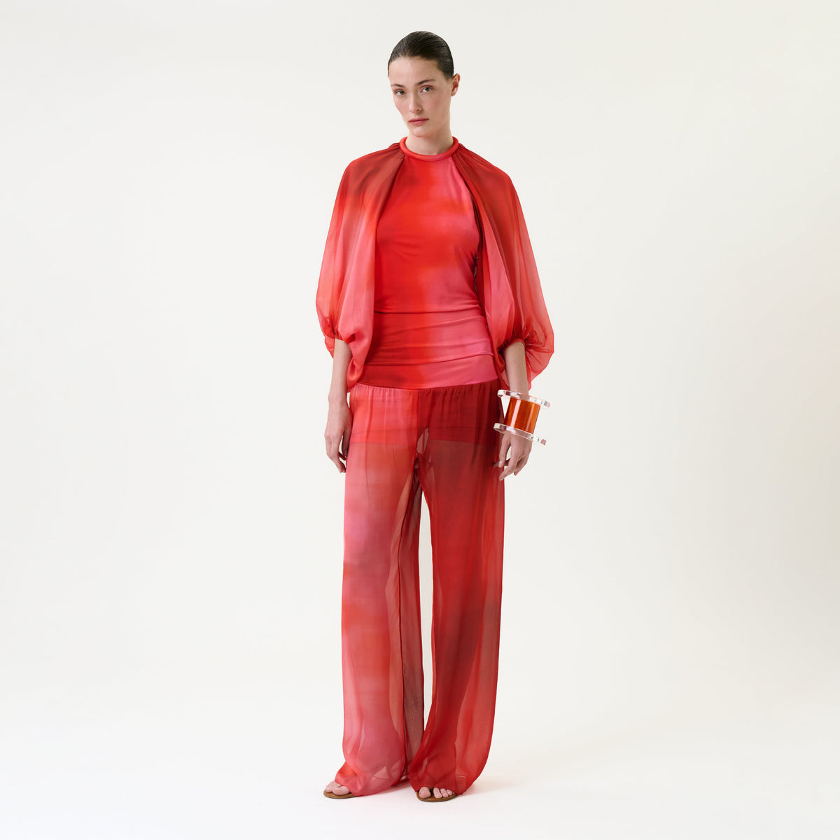 A woman stands against a white background wearing the sheer red Lene Pant paired with a fitted top and matching cape. She holds an orange clutch and wears flat sandals, with her hair slicked back.