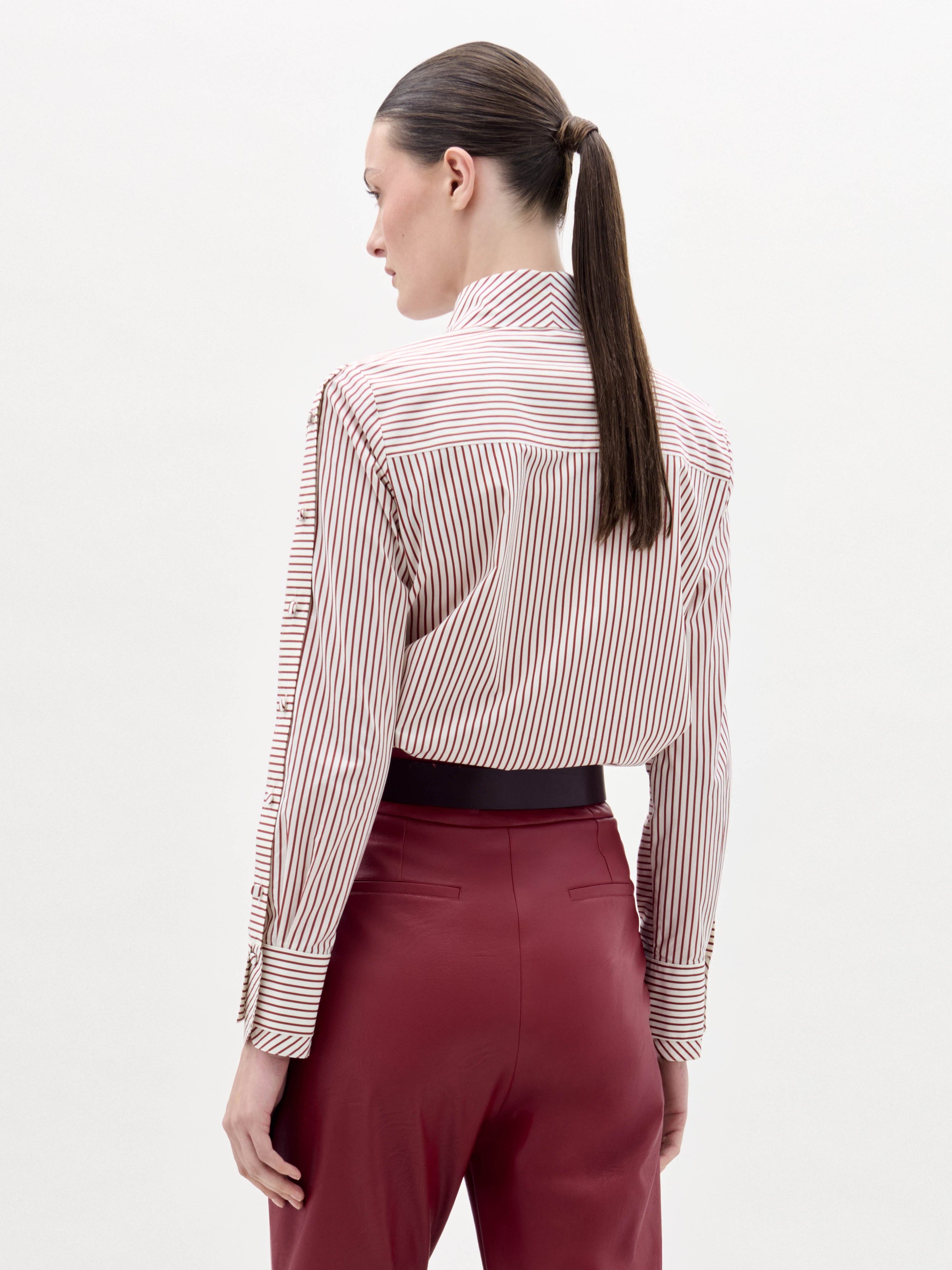 A woman stands against a plain background wearing the Premium Blouse, featuring stripes and a large red floral design, paired with red belted trousers and a decorative belt buckle.