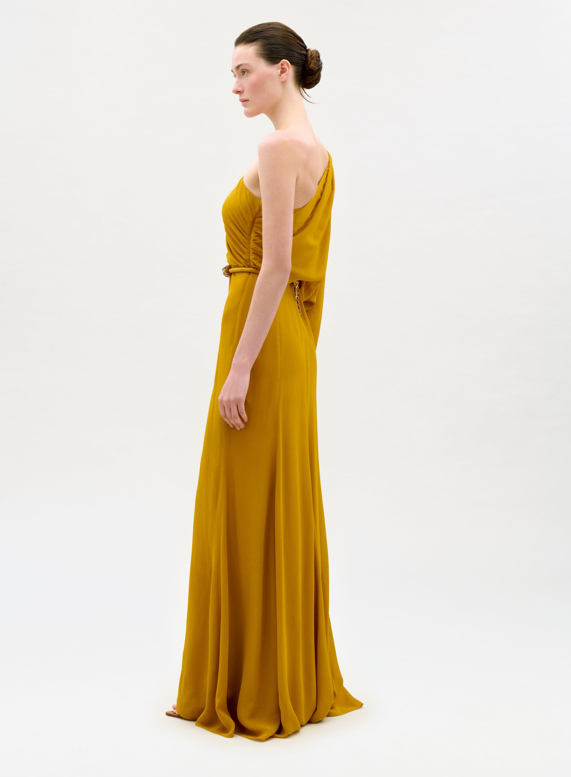 A woman in the Minerva Dress, a mustard yellow one-shoulder floor-length gown, stands in profile against a plain light background with her hair styled in a neat bun.