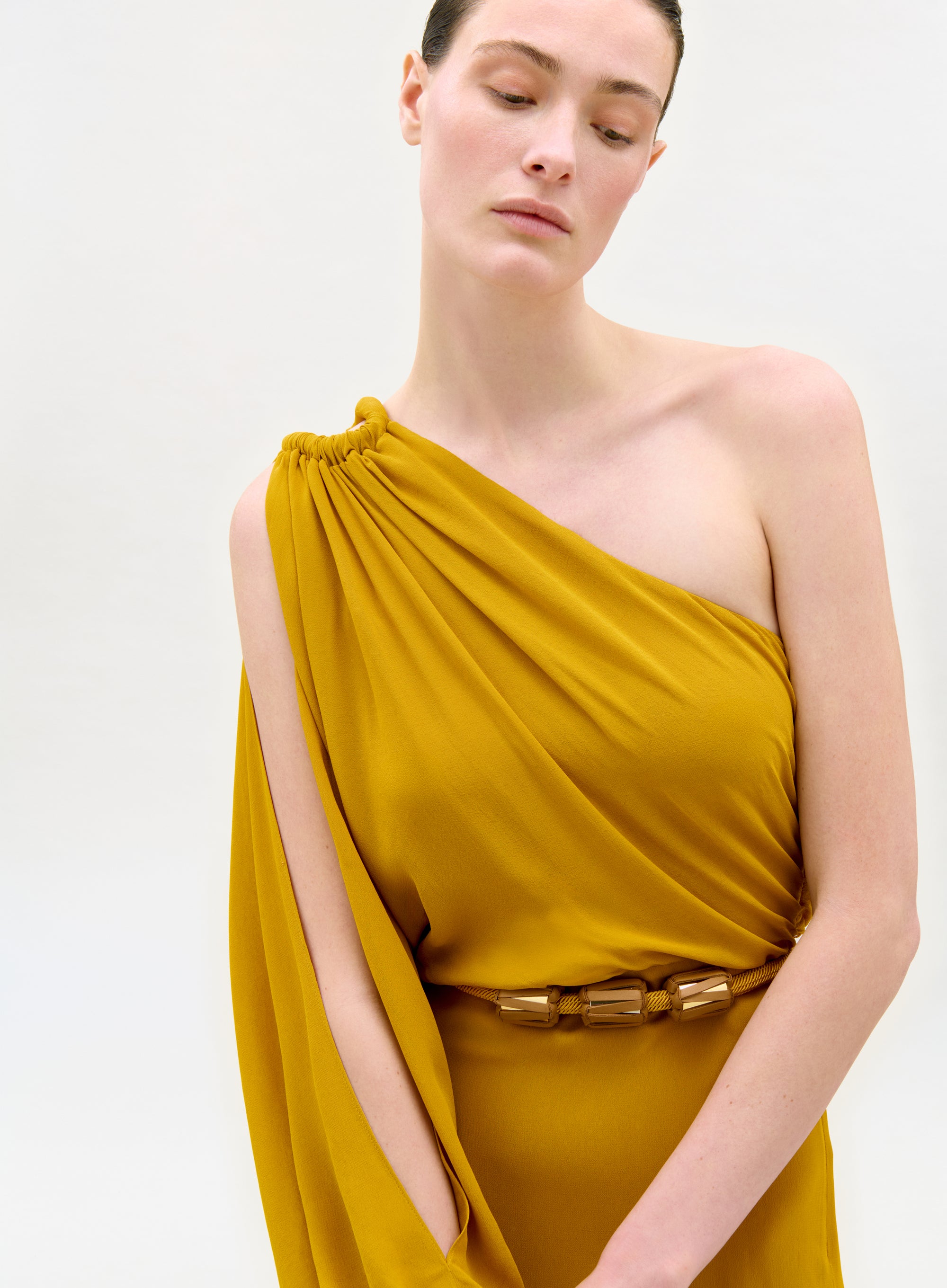 A woman stands serenely against a plain light background, wearing the Minerva Dress—mustard yellow, one-shoulder, draped with gathered shoulder detail and a chic gold belt.