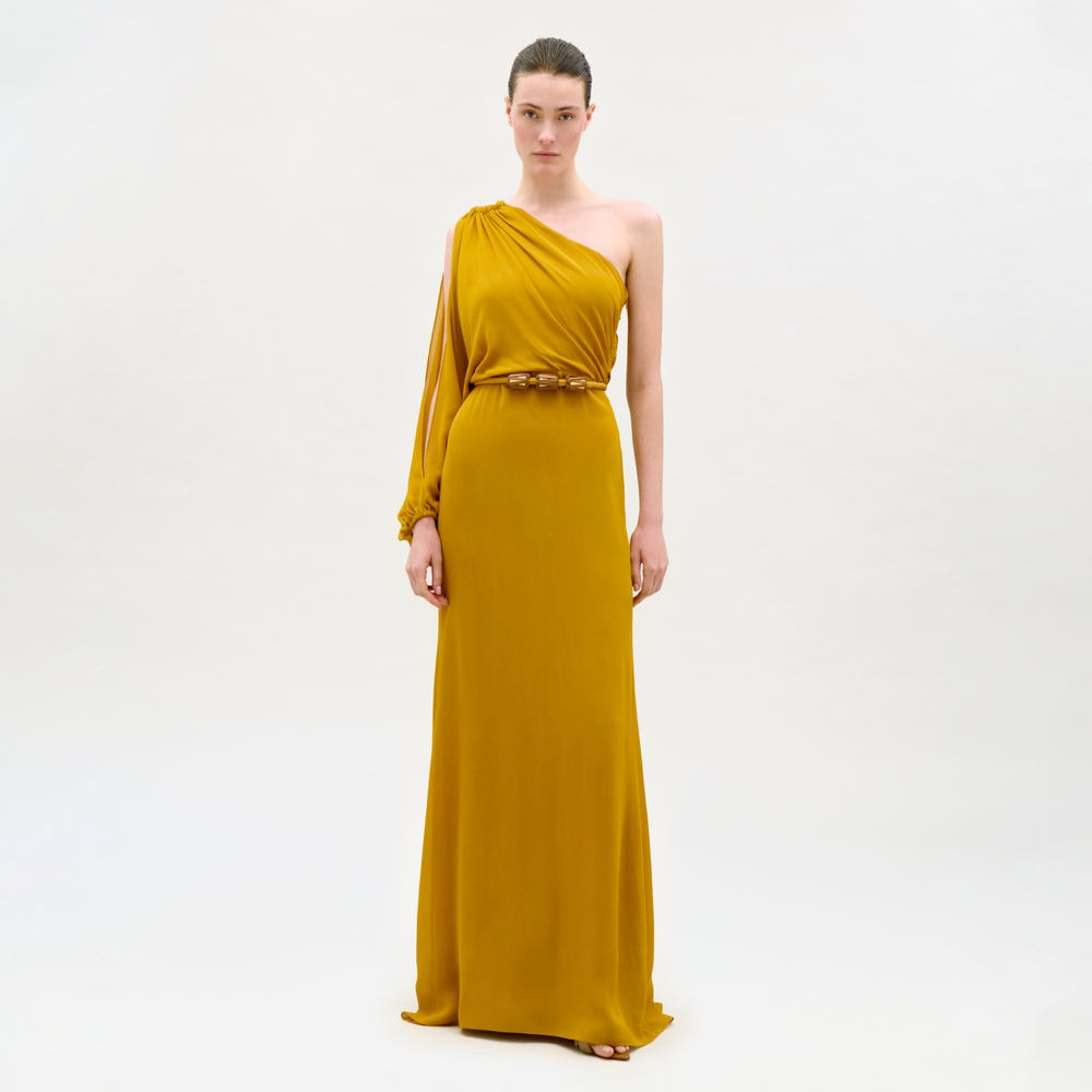A woman stands against a plain white background wearing the Minerva Dress, a mustard yellow, floor-length, one-shoulder gown with a belted waist.