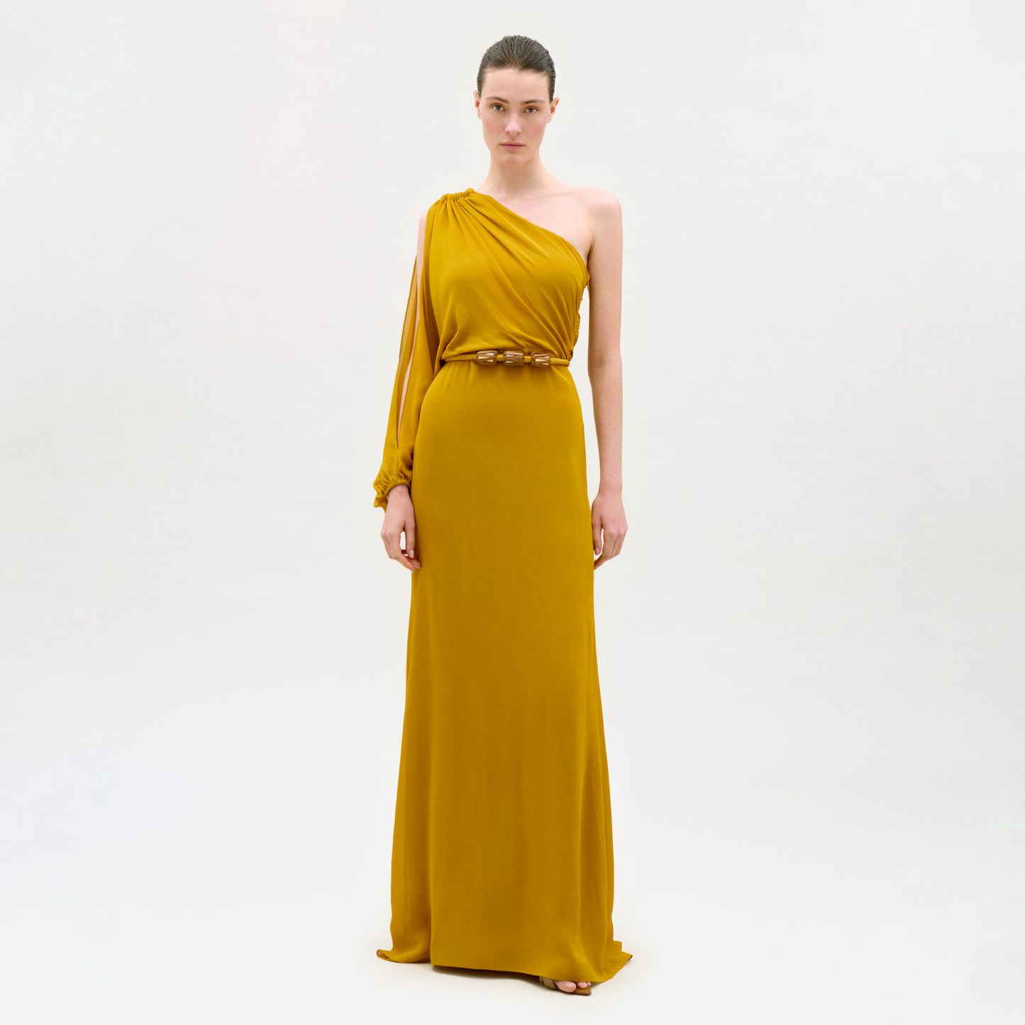 A woman stands against a plain white background wearing the Minerva Dress, a mustard yellow, floor-length, one-shoulder gown with a belted waist.