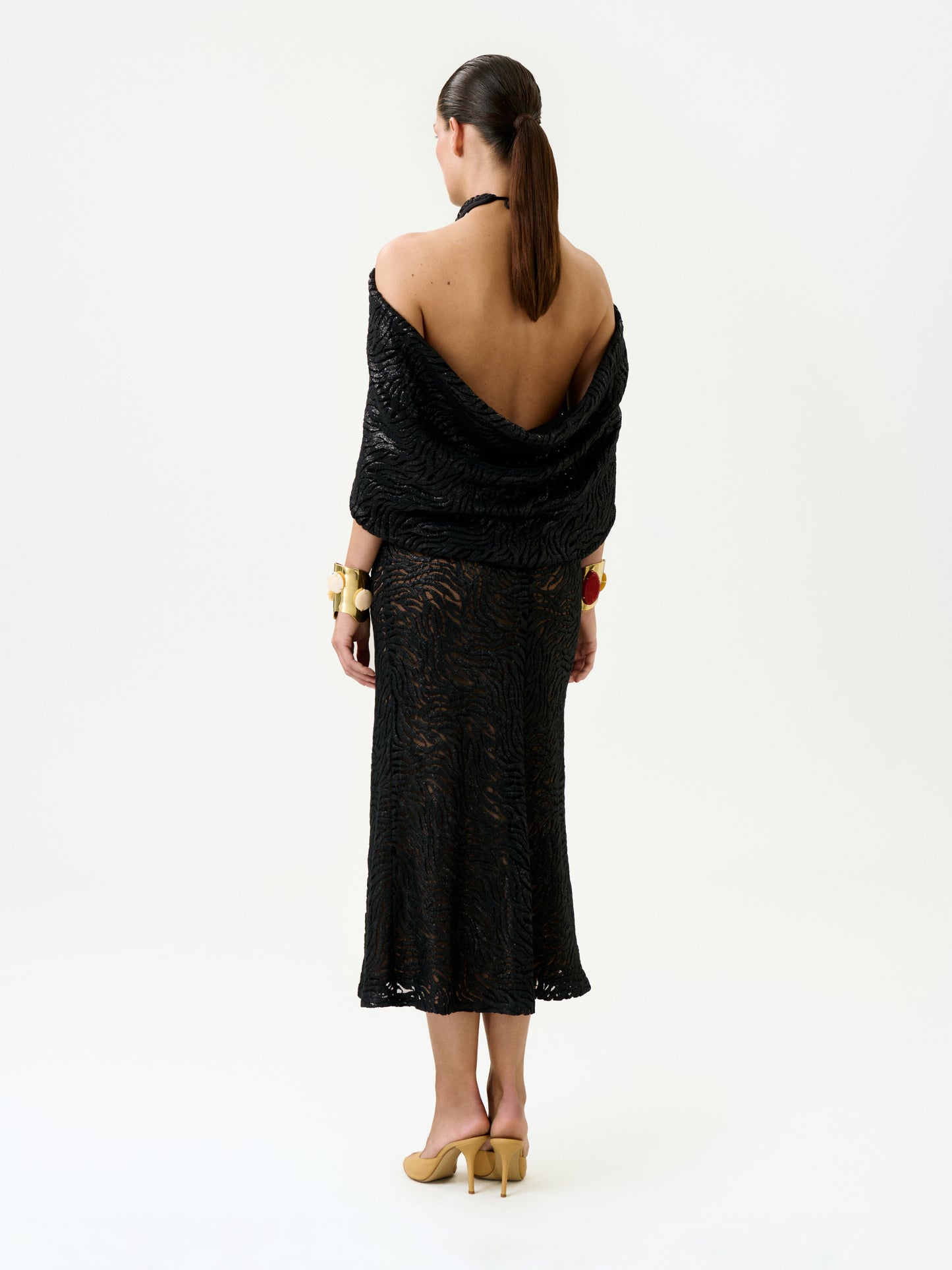 A woman with a low ponytail and chunky bracelets stands against a white background, facing away and wearing the Neda Dress—a black textured, backless midi dress made from Italian fabric—with a shawl and beige heels.