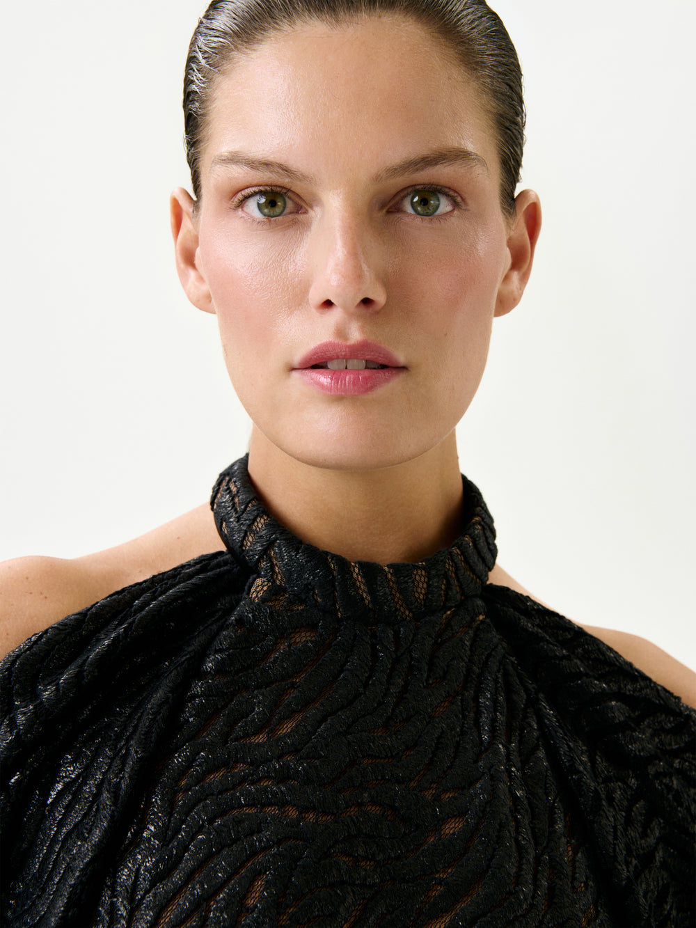 A person with slicked-back hair wears the Neda Dress—a textured black high-neck piece crafted from Italian fabric—while looking directly at the camera against a plain, light background.