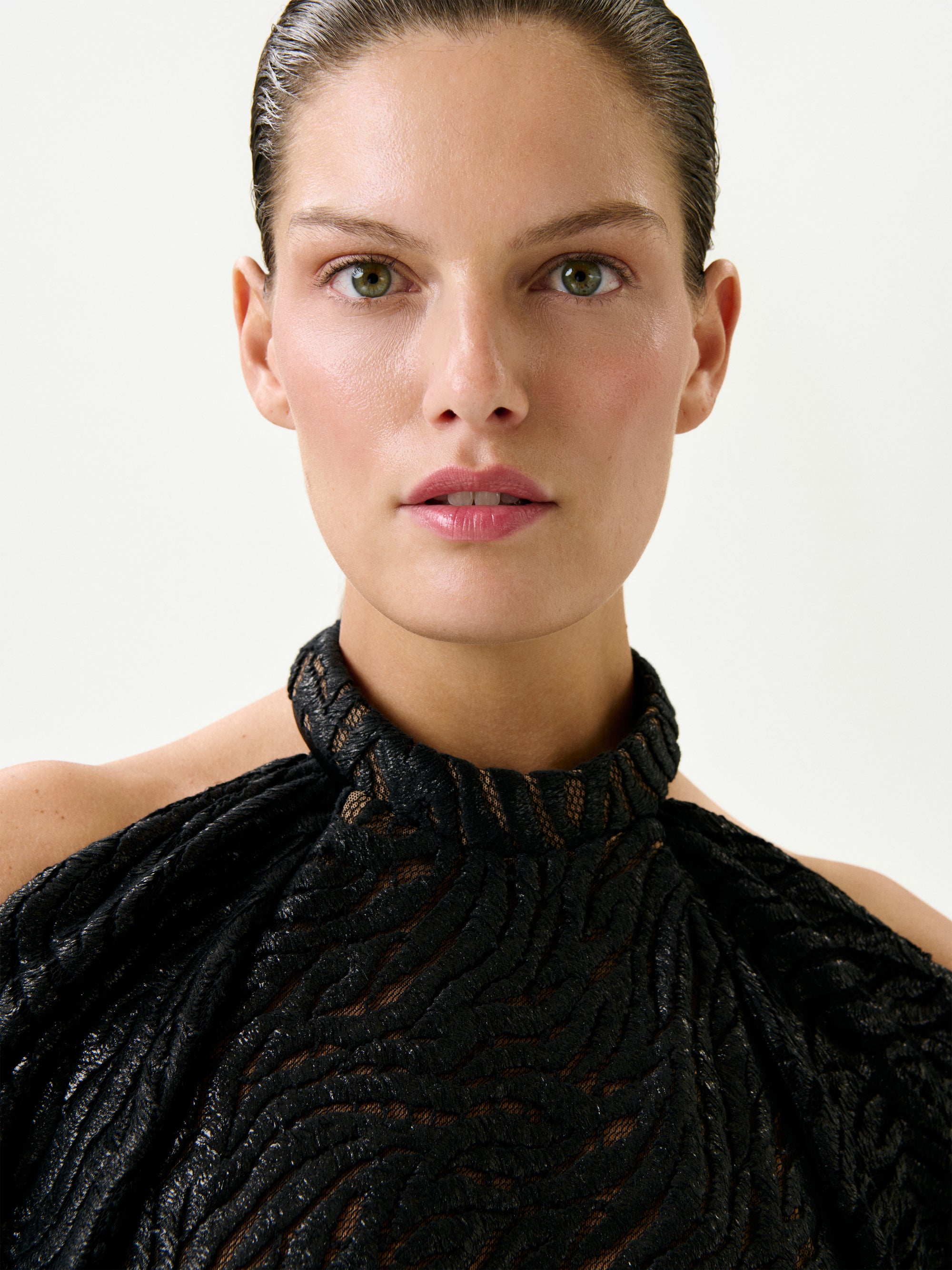 A person with slicked-back hair wears the Neda Dress—a textured black high-neck piece crafted from Italian fabric—while looking directly at the camera against a plain, light background.